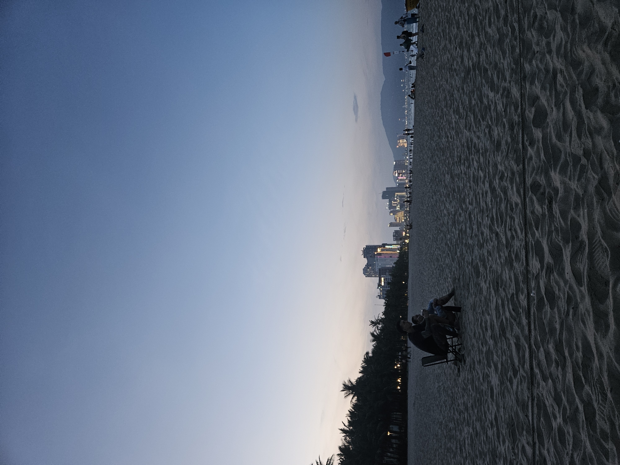The image depicts a serene beach scene at dusk in Đà Nẵng, Vietnam. The sky transitions from a deep blue at the top to a pale orange and pink near the horizon, indicating the sun has recently set. In the distance, a city skyline with illuminated buildings stands against a hazy mountain range. Closer to the shore, a line of palm trees forms a dark silhouette against the twilight sky, with some lights visible through the foliage.
On the sandy beach, several people are visible, including a couple sitting in chairs, looking out towards the ocean and city. The sand is textured with ripples and footprints, suggesting it is a well-trodden area. Further down the beach, other figures can be seen walking and standing, some silhouetted against the fading light. The overall atmosphere is peaceful and relaxed, capturing the end of a day by the sea. There is no visible text in the image.