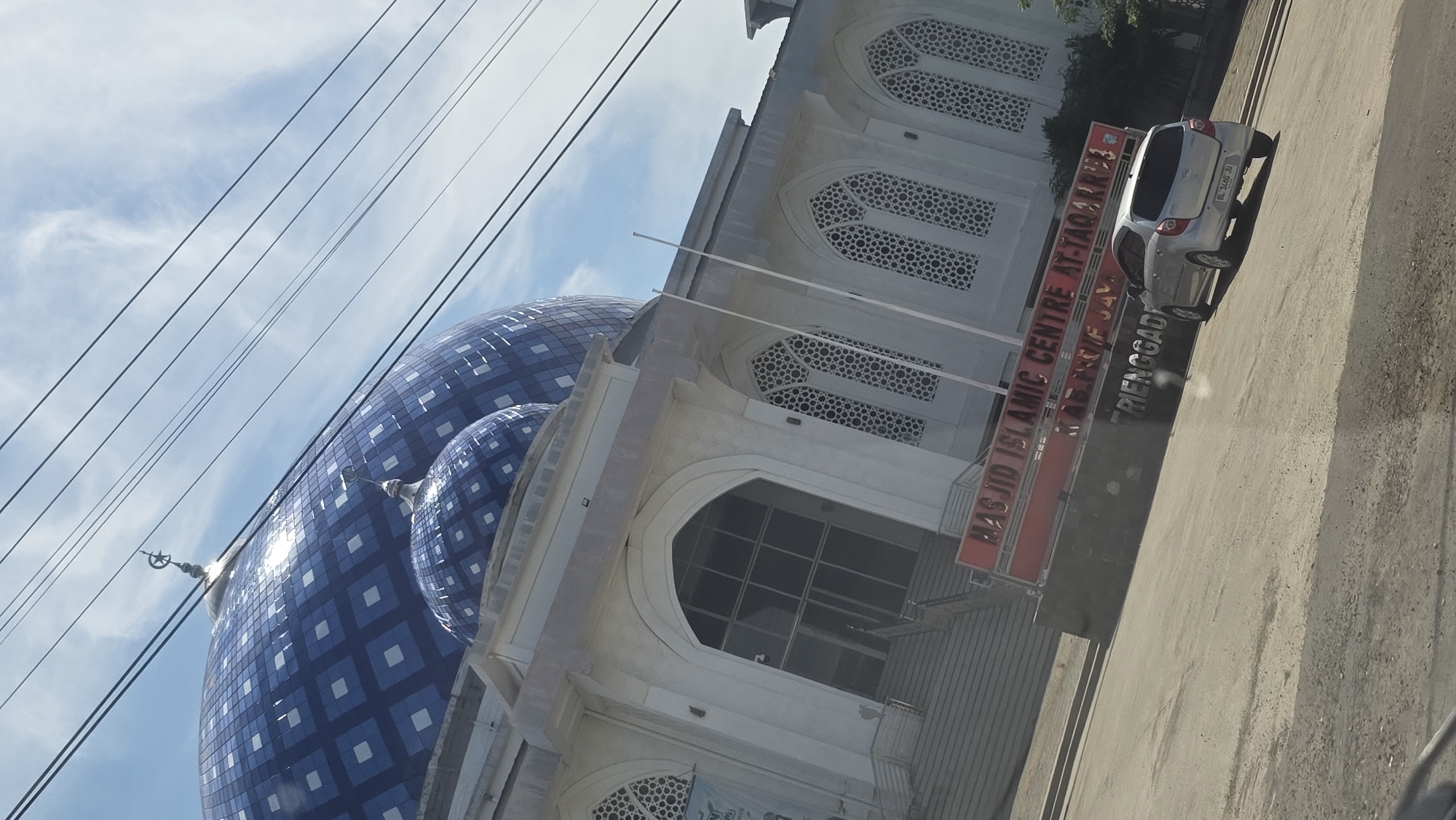 The image shows the Masjid Islamic Centre At-Taqarrub in Paya Tringgadeng, Aceh, Indonesia. The mosque features a large blue dome with a white geometric pattern. A silver car is parked in front of the mosque on the road. A sign reading "MASJID ISLAMIC CENTRE AT-TAQARRUB, KUALA IDIE, ACEH TIMUR, PAYA TRIENGGADENG" is visible. Overhead power lines are present in the top left of the frame. The sky is blue with scattered clouds.