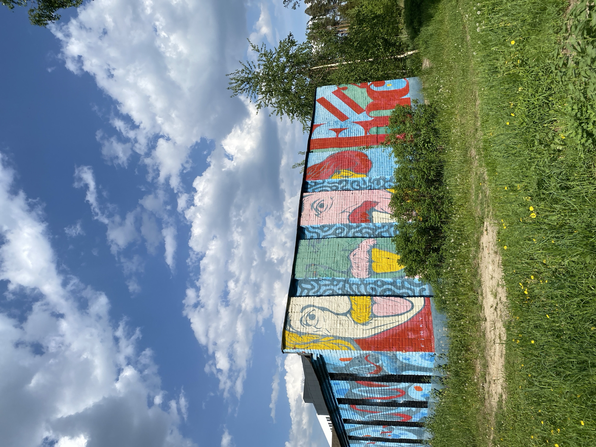 The image shows a section of a building's exterior wall with a mural painted on it. The mural features abstract and colorful depictions of faces and possibly text. The building is located next to a grassy area with a dirt path running alongside it. The sky is visible in the background, filled with white clouds against a blue sky. The specific location within the city of Unknown, Unknown, cannot be determined from this image alone.