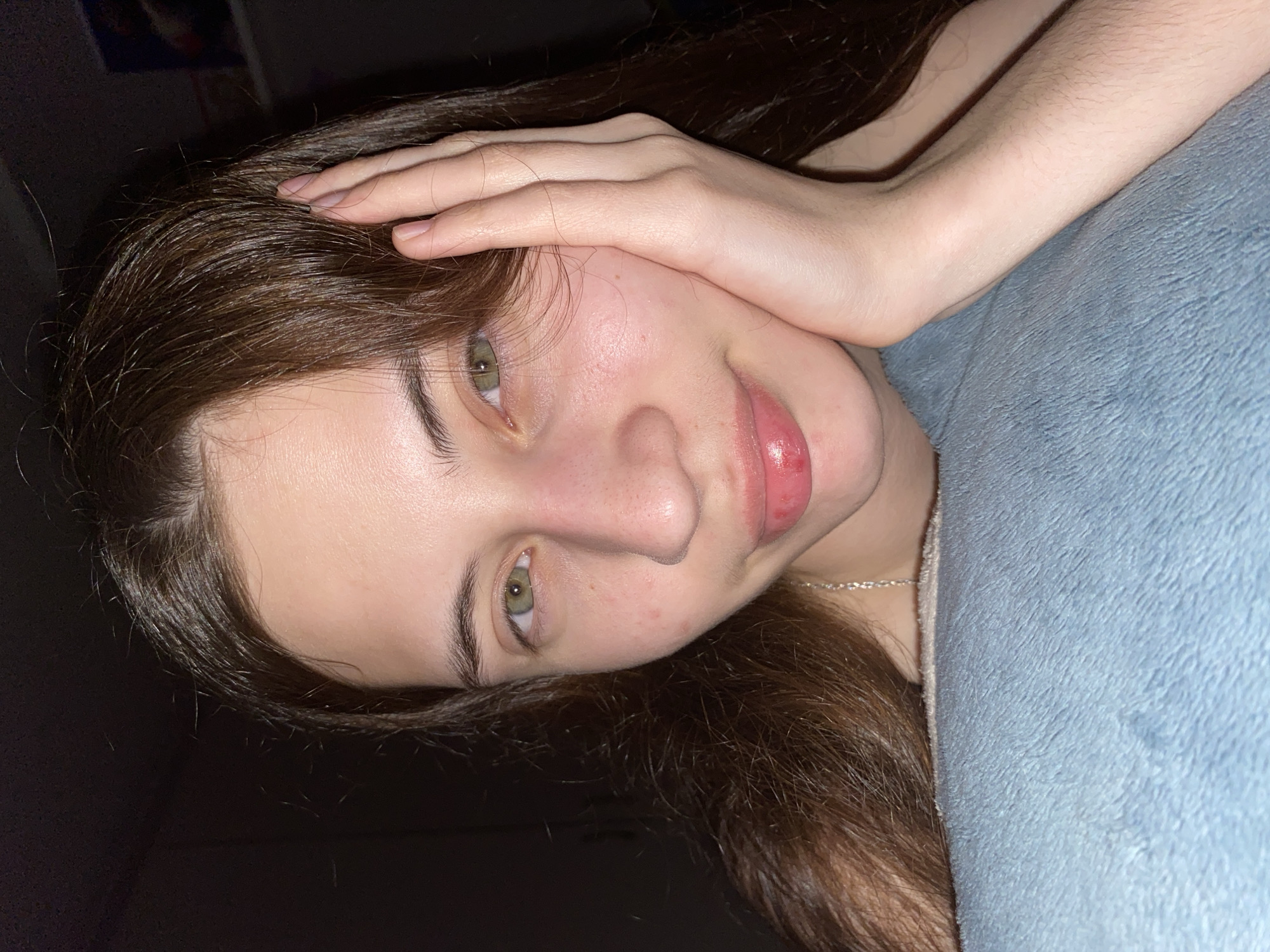 A young woman with long brown hair is lying on a light blue soft surface, possibly a blanket or pillow. She has fair skin, green eyes, and pink lips. Her right hand is resting on her forehead. A silver necklace is visible around her neck. The background is dark. The photo's angle suggests it was taken from above, possibly with a phone.
There is no indication of the location being Votkinsk, Russia in the image.