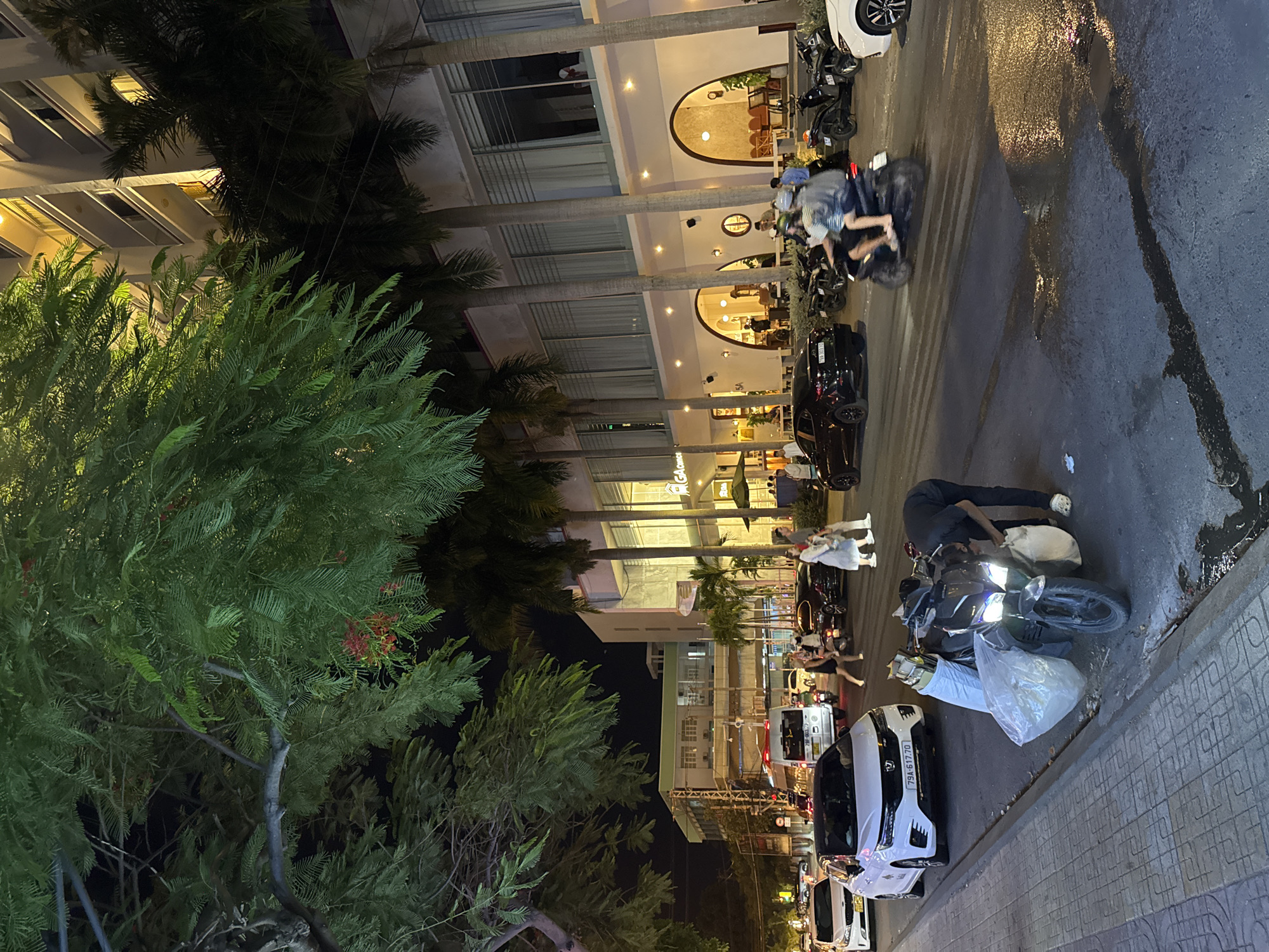 A nighttime street scene in Nha Trang, Vietnam, shows a busy road with traffic and pedestrians. A man is interacting with a scooter loaded with plastic bags, positioned on the side of the road. Several cars and motorcycles are on the street, and pedestrians are walking on the sidewalk and crossing the road. The background features a building with arched entrances and palm trees, illuminated by interior and exterior lighting. A large tree with green foliage dominates the left side of the frame.
