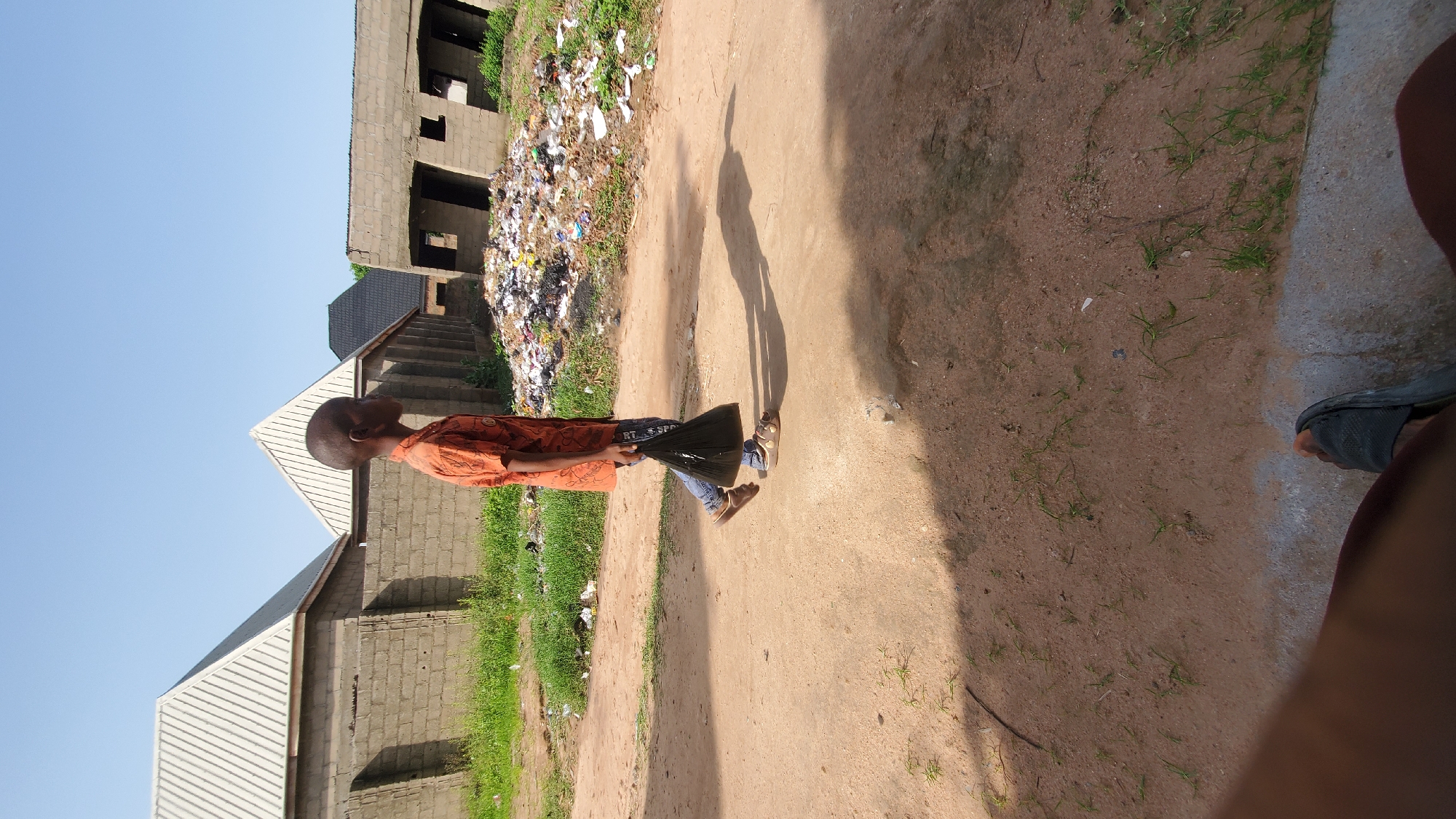 A young boy walks down a dirt path in Jalingo, Nigeria, carrying a black plastic bag in his right hand. He is wearing an orange patterned shirt and blue jeans. To his left are several unfinished brick buildings with corrugated metal roofs, and to his right is a large pile of garbage. The sky is clear and blue.