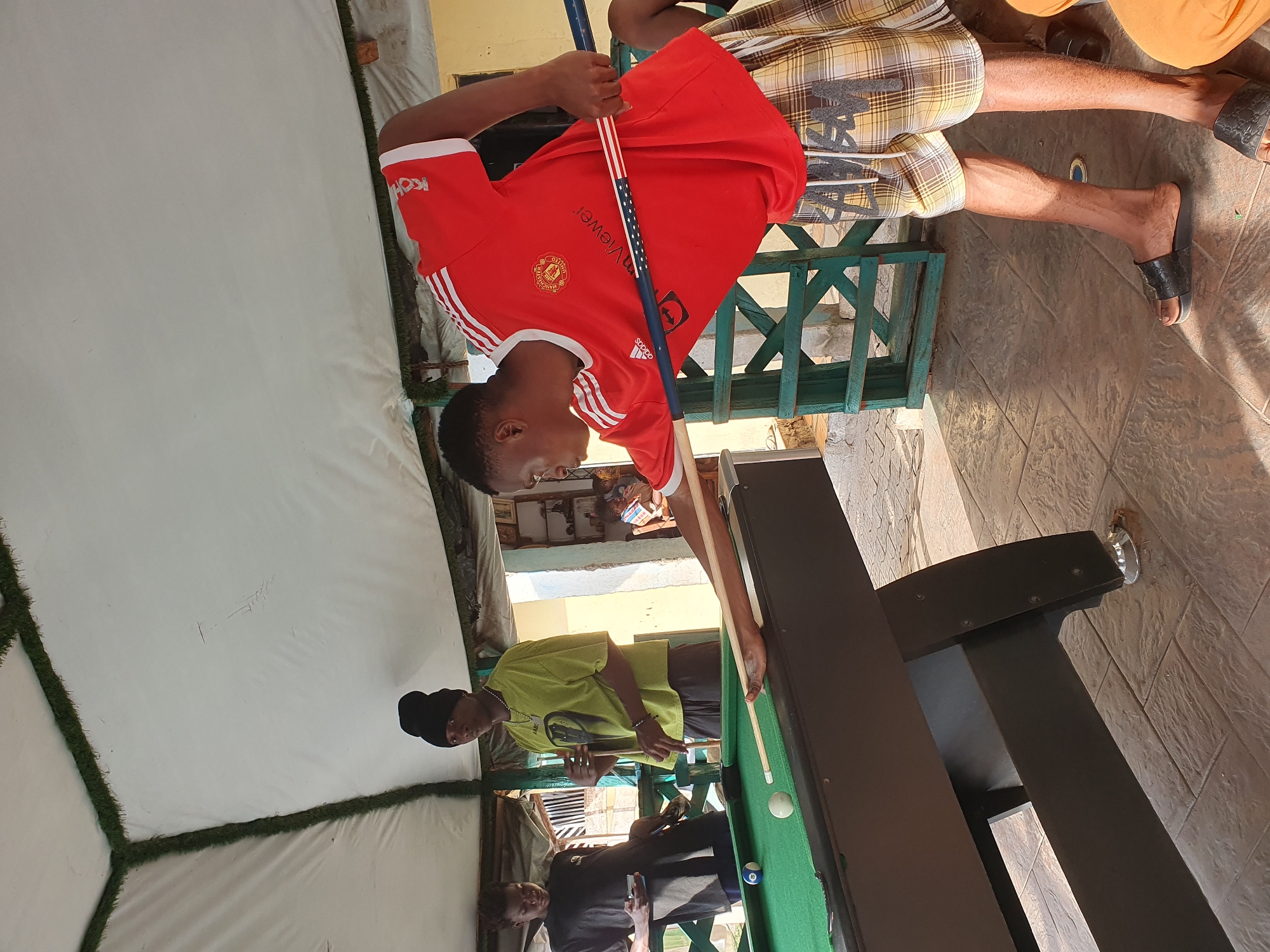 In Lokoja, Nigeria, three young Black men are engaged in a game of pool. The main subject, wearing a red Manchester United jersey and plaid shorts, is in the foreground, actively playing with a cue stick. To his left, another man in a bright green shirt and black beanie holds a cue stick, looking towards the pool table. A third individual in a black shirt stands further left, holding a smartphone. The scene is set outdoors, under a white tent structure with green trim, and a green fence is visible behind the players. The ground is paved with textured tiles, and a pool table with green felt and billiard balls is the central focus. Sunlight casts shadows, indicating it is daytime.