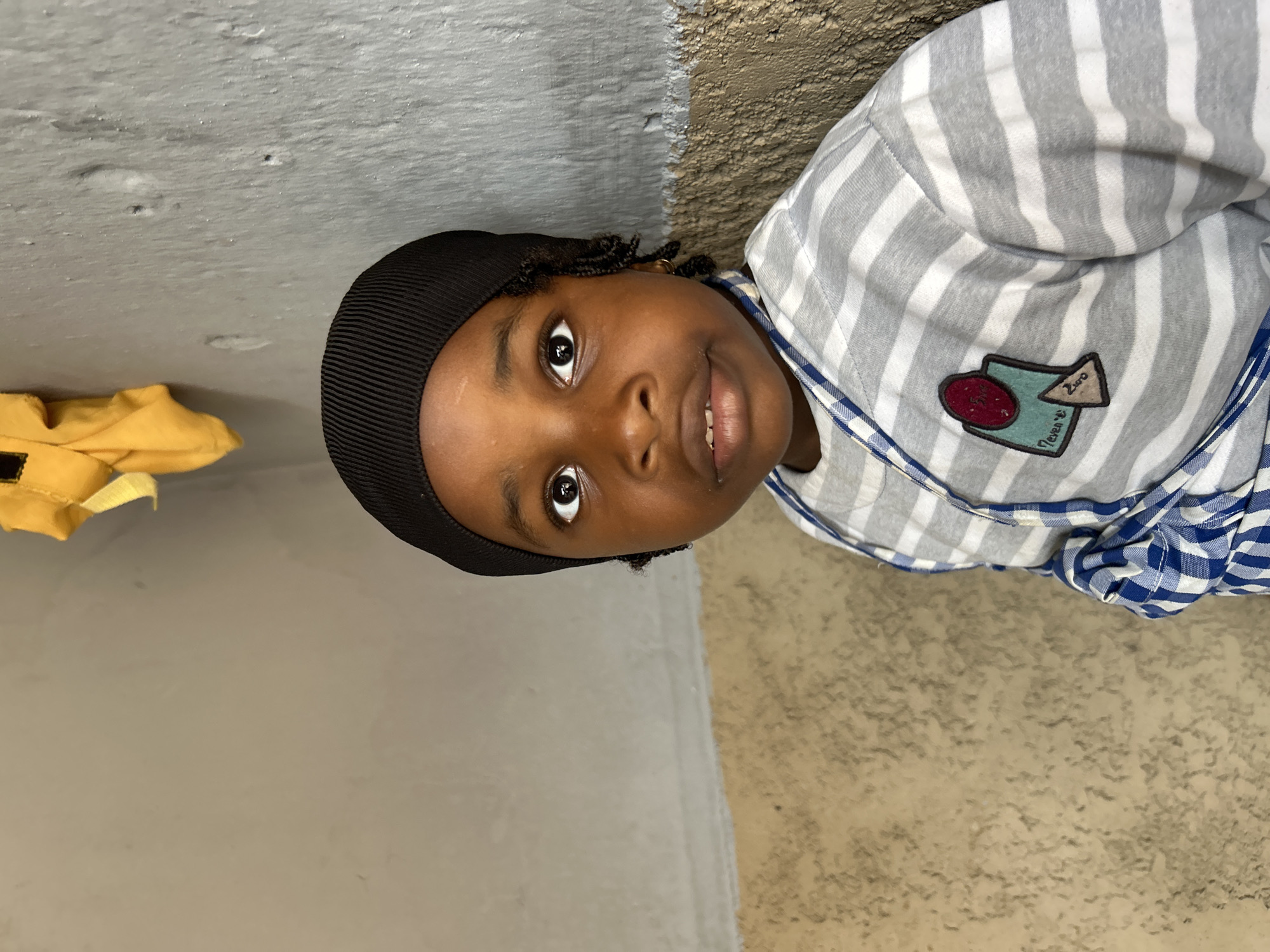 A close-up, tilted portrait of a young child is presented. The child has dark skin, brown eyes, and is wearing a black knit cap. The child is wearing a striped shirt with a patch displaying an abstract design with text. The background appears to be a wall with a color transition, one side is gray and the other is a textured tan color. A piece of yellow fabric is visible on the left side of the frame. The location of the photo is unknown, including whether it was taken in the city of Jos, Nigeria.