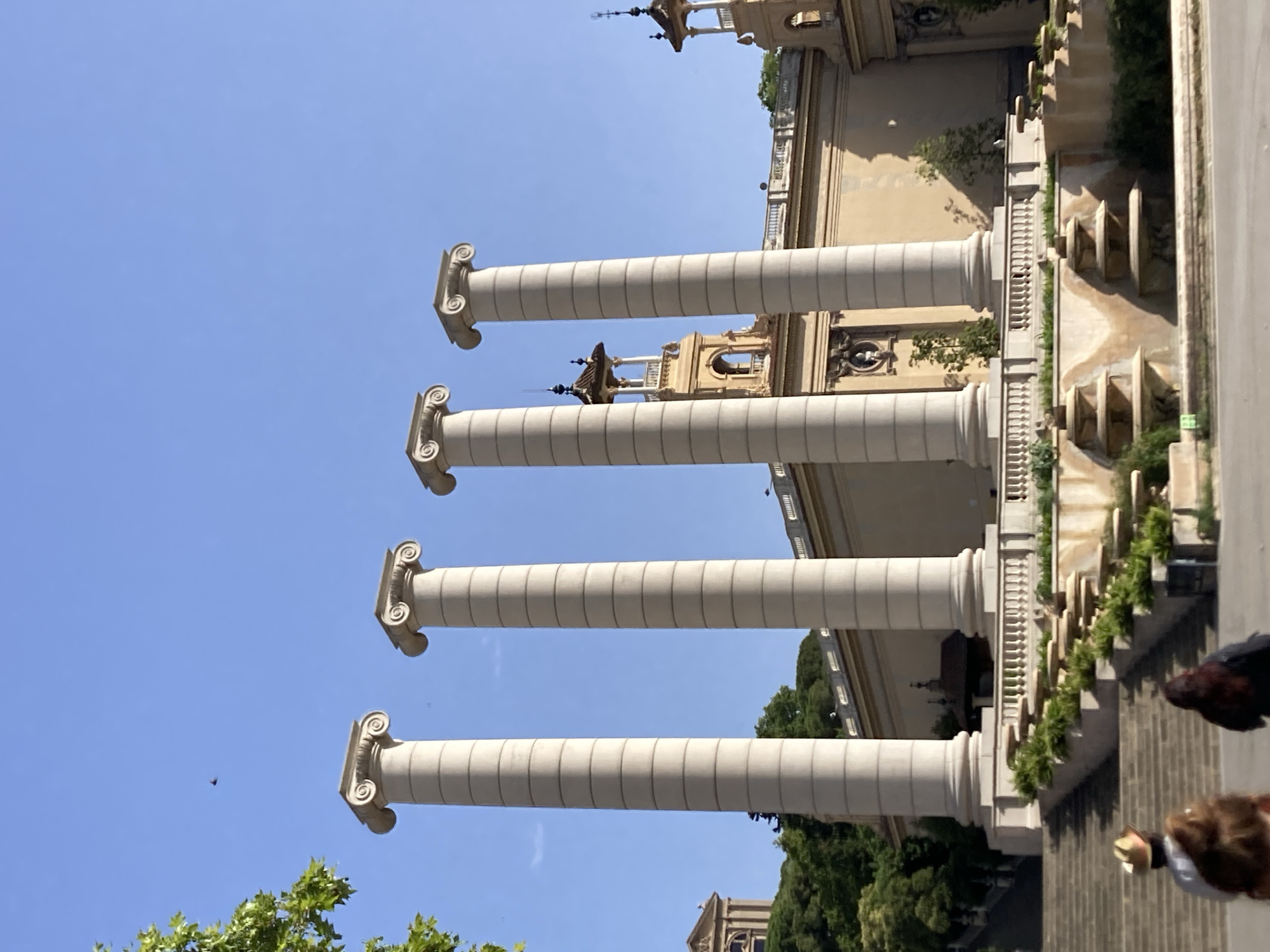 The image depicts the Four Columns of Montjuïc in Barcelona, Spain, under a clear blue sky. Four large, fluted Ionic columns stand prominently in the foreground, their capitals featuring volutes. Behind the columns, a long, light-colored building with arched windows and decorative elements stretches across the frame. A tower with an ornate roof is visible behind the second and third columns from the left. Green trees and foliage are present on a hillside to the left of the building. In the lower portion of the image, a terraced fountain with multiple tiers and lush green plants is situated in front of a white balustrade. A wide stone staircase leads up towards the fountain area. Two individuals, their backs to the camera, are partially visible on the lower steps of the staircase, one wearing a hat. The scene is bathed in sunlight, casting shadows on the building and ground.