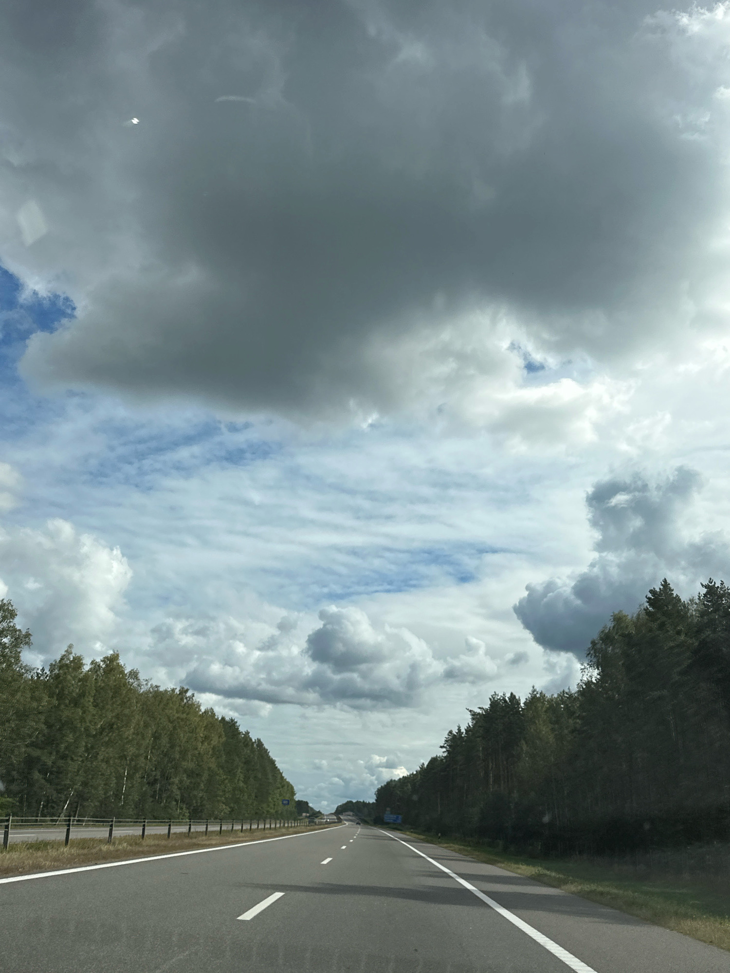 The image shows a highway in the city of Пухавіцкі сельскі Савет, Belarus, on a cloudy day. The highway is asphalt and has white lane markings. On the sides of the highway are trees, mostly deciduous on the left and coniferous on the right. The sky is filled with dark, heavy clouds, with patches of blue sky visible. There is a white line of a guardrail visible on the left side of the road. The perspective is from a vehicle traveling on the highway.