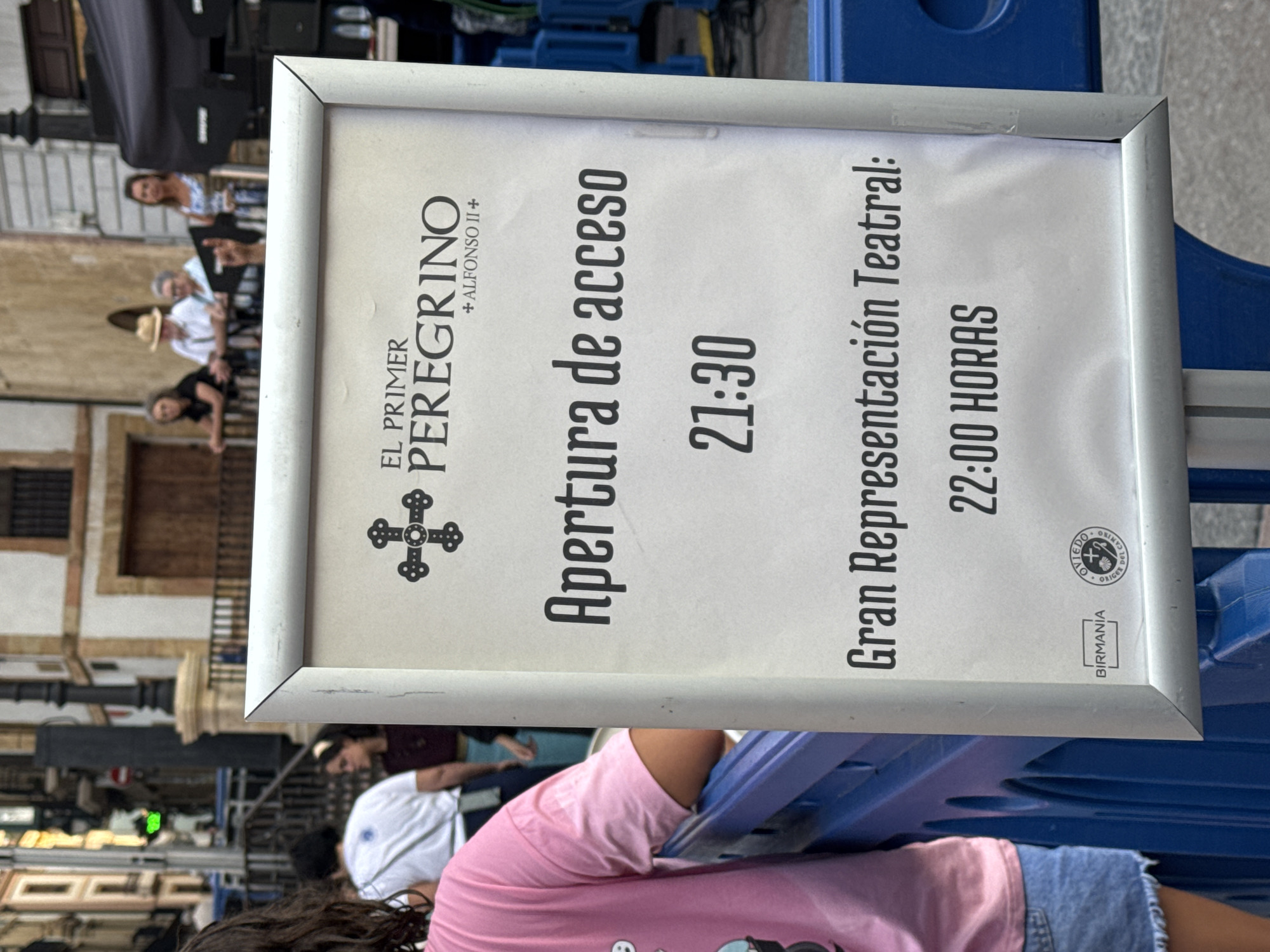 The image shows a sign in Oviedo, Spain. The sign is framed and displays text related to an event. The text includes "EL PRIMER PEREGRINO + ALFONSO II+", "Apertura de acceso 21:30", and "Gran Representación Teatral: 22:00 HORAS". Below this, there are two logos, one reads "BIRMANIA" and the other is a circular emblem with "OVIEDO ORIGEN DEL CAMINO" written around it. In the foreground, the shoulder and arm of a person wearing a pink t-shirt and denim shorts are visible. In the background, several people are standing, and part of a building with architectural details can be seen. The environment appears to be an outdoor public space with some barriers.