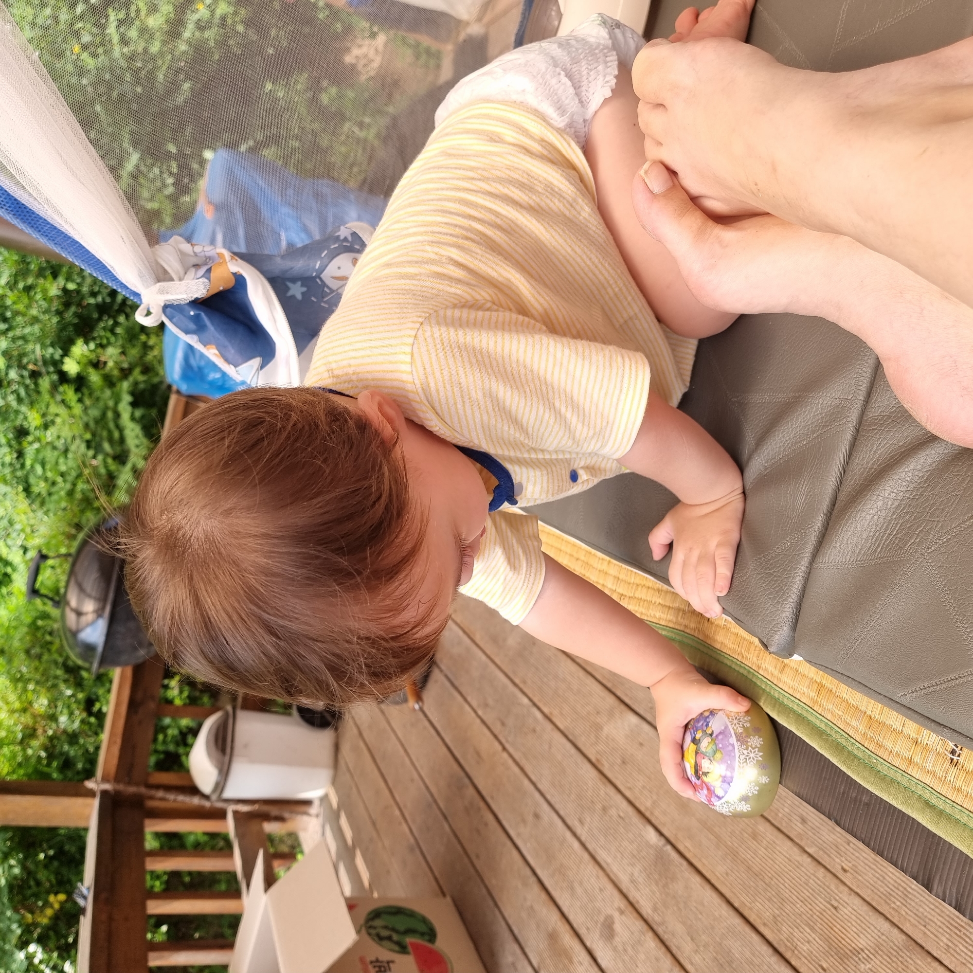 A toddler in a yellow and white striped t-shirt and a diaper is playing with a small, round toy on a wooden deck. The toddler is sitting on a grey cushion, with their legs bent. In the foreground, a person's feet are visible. In the background, there is a barbecue grill and lush green foliage. The location is identified as Dunnae-myeon, South Korea.