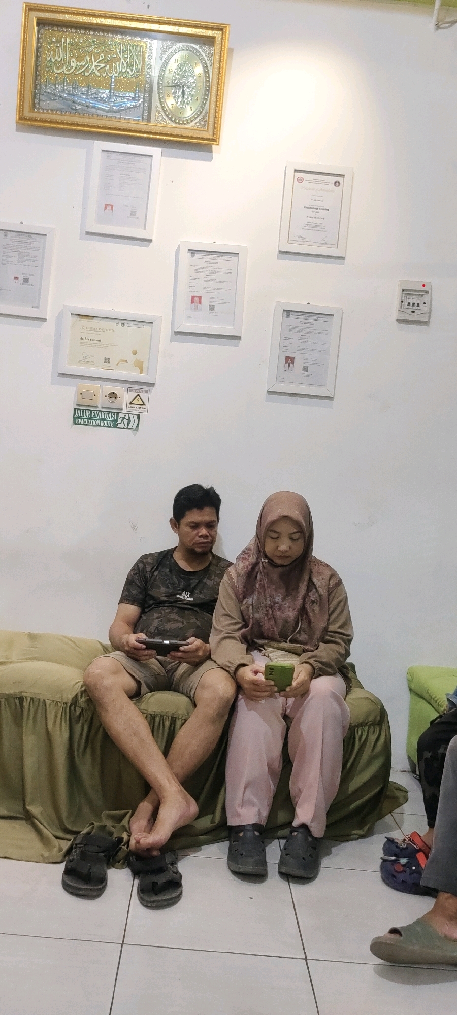 The image depicts an indoor scene, likely a living room or waiting area, in Sungai Sipai, Indonesia. A man and a woman are seated on a green sofa. The man, wearing a camouflage t-shirt and shorts, is holding a dark-colored electronic device, possibly a tablet or phone. The woman, dressed in a patterned hijab and pink pants, is looking down at a green smartphone. Their feet, bare and with crossed legs, are visible in the foreground. Several pairs of black sandals are placed on the tiled floor near the sofa.
The wall behind them is adorned with framed documents and a decorative piece that includes a clock and what appears to be Islamic calligraphy. A framed "EVACUATION ROUTE" sign is also visible on the wall, along with electrical outlets and a light switch. The overall environment appears to be residential or a small office space, with the white walls and various framed items suggesting a sense of order and personalization. The lighting is bright, indicating an indoor setting with artificial light sources.