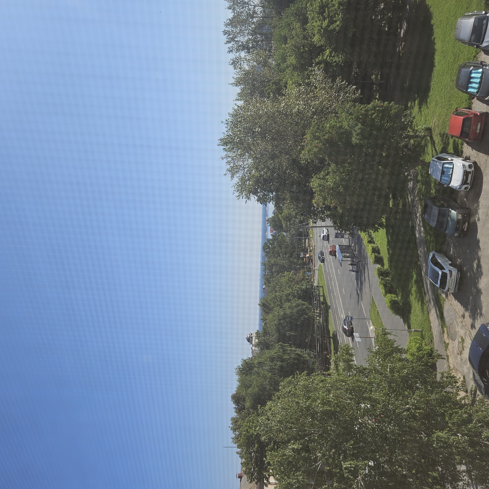 The image depicts a city scene from an elevated perspective, likely a residential building, overlooking a street and a park area. The sky is clear blue, with no clouds visible. A wide, multi-lane road runs horizontally across the mid-section of the image, with several cars driving on it. On the right side of the road, there is a bus stop shelter with a few people standing underneath. The road is bordered by green grass and trees, with a sidewalk next to it.
In the foreground, on the right side, several cars are parked in a lot. From left to right, a white SUV, a dark green SUV, and a white sedan are clearly visible. Further to the right, partially obscured by trees, are a red car, a dark gray car, and a silver car. On the left side of the image, closer to the foreground, a dark blue car is parked.
Behind the road and the park, a dense line of green trees stretches across the image. In the distance, beyond the trees, a body of water, likely a river or lake, is visible, with a city skyline on the horizon. A domed building is prominent on the skyline, suggesting a significant landmark.
Based on the presence of a domed building and the body of water, and cross-referencing with typical landmarks of Cheboksary, Russia, the image is likely taken from a location overlooking the Volga River, near the city center. The architecture and vegetation are consistent with a Russian urban environment. The overall scene suggests a sunny day in a city with significant green spaces.
