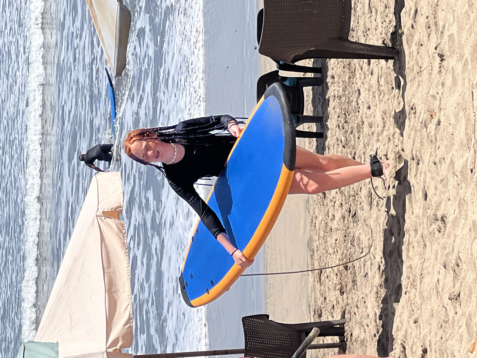 A young woman with auburn braided hair stands on a sandy beach in Kuta, Indonesia, holding a blue and orange surfboard. She is wearing a black long-sleeved shirt and a white shell necklace. Her legs are bare, and she has a black ankle leash attached to her right foot, which connects to the surfboard. In the background, the ocean waves are breaking, and other people are visible in the water, one of whom is in the process of surfing. Two beach umbrellas and several empty plastic chairs are also present on the sand. The sun is shining brightly, casting shadows on the beach.