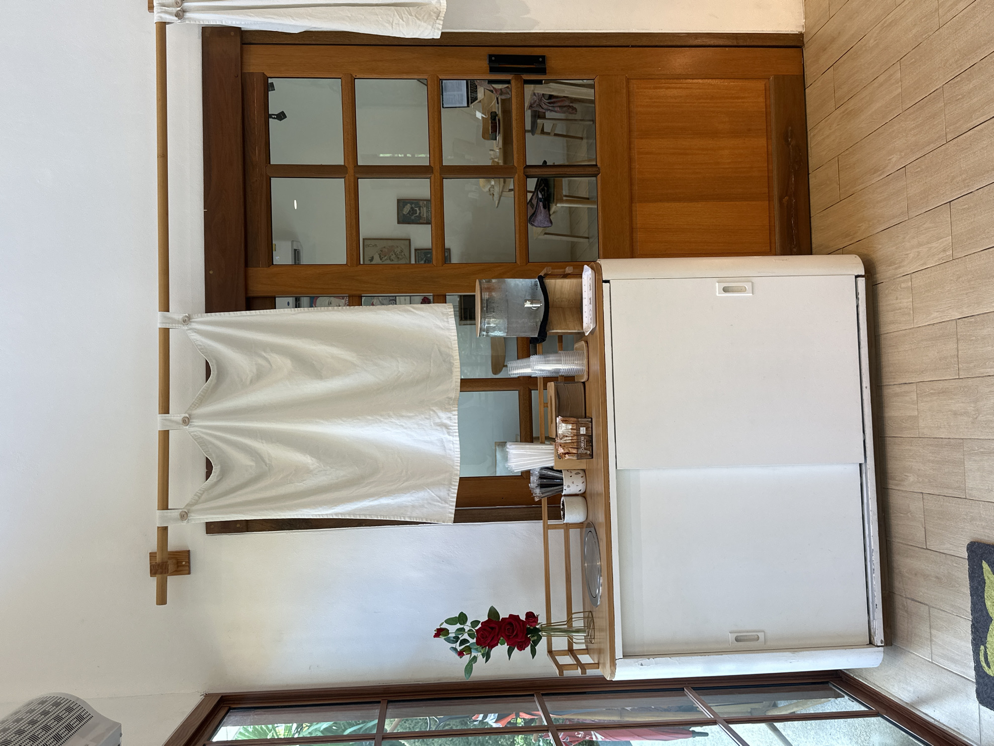 This image depicts an indoor scene, likely a cafe or restaurant counter area, in Kathu, Thailand. A white cabinet with sliding doors is positioned against a white wall. A wooden countertop sits atop the cabinet, adorned with various items. These include a vase of red roses, cups, straws, napkins, and a large glass water dispenser. To the right of the counter is a wooden door with glass panes, revealing a glimpse of tables, chairs, and a laptop inside the adjacent room. The floor is tiled with light brown wooden planks. A white curtain hangs from a wooden rod above the counter and door, partially obscuring the view. The lighting suggests a well-lit indoor environment, possibly during the day.