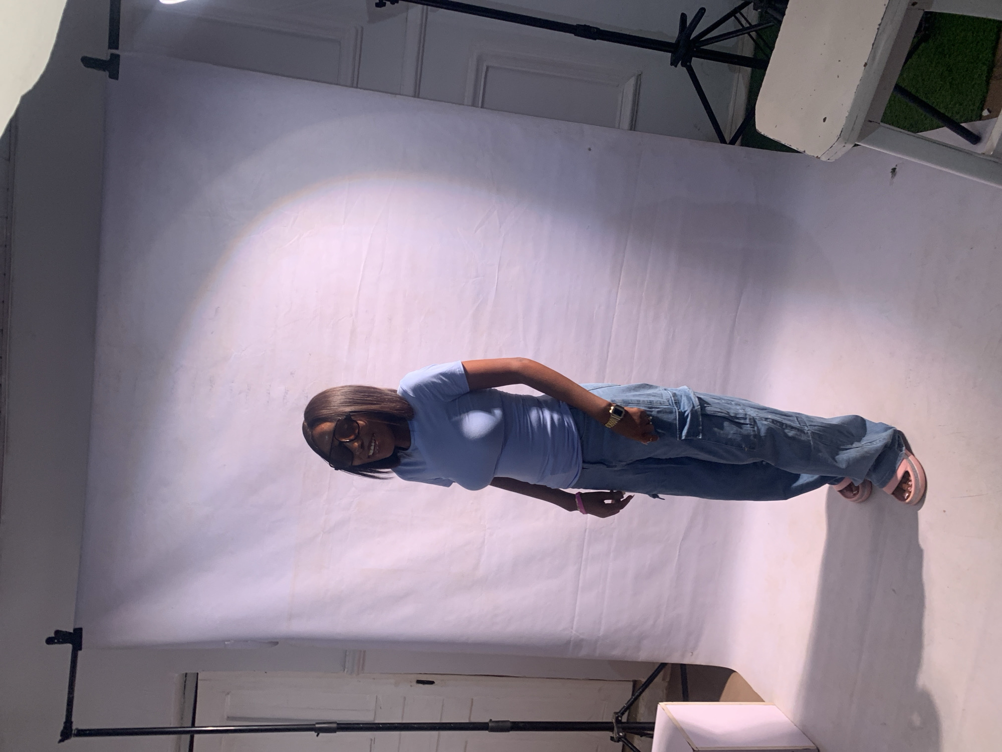 The image depicts a woman standing against a white backdrop. She is wearing a light blue T-shirt, light blue jeans, and pink sandals. She also sports dark sunglasses and a gold watch. The setting appears to be a photography studio or a similar space designed for capturing images. Studio lighting equipment is visible, including a spotlight and stands. The specific location is unknown and cannot be confirmed as Jos, Nigeria.