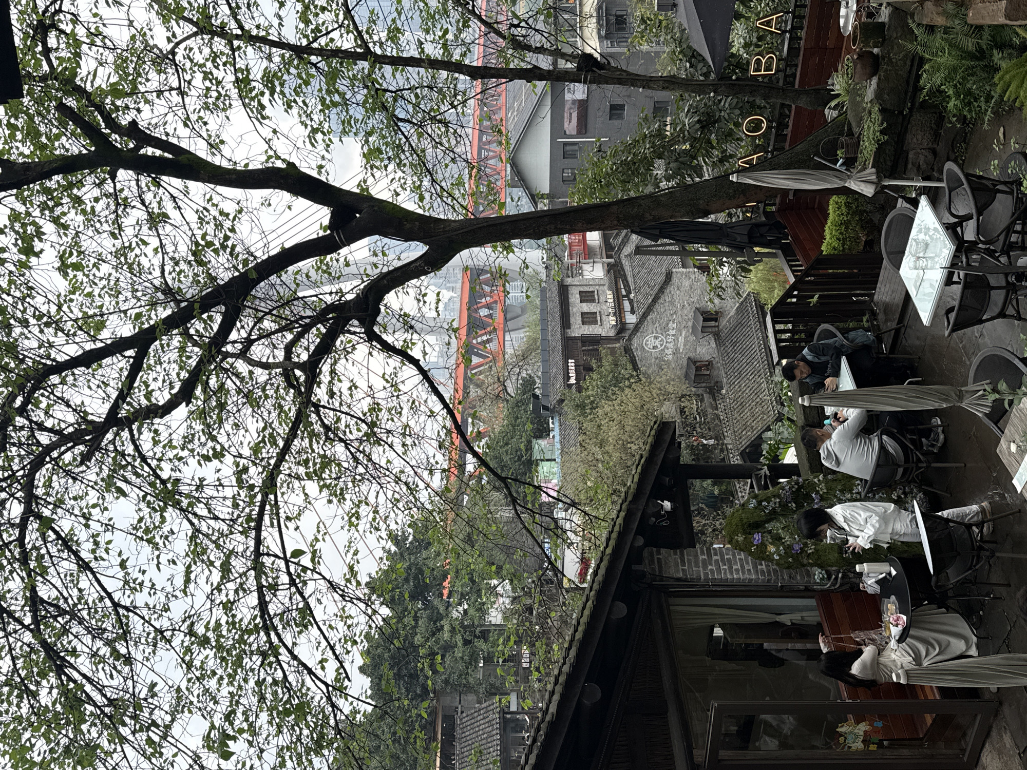 The image displays an outdoor cafe terrace located on a hillside in Nan'an District, China, under an overcast sky. The ground surface of the terrace appears damp.
In the left foreground, a section of a building with a dark, sloped tiled roof, a brick facade, and large glass doors/windows is visible. Two women are positioned near a round, dark-topped table. One woman, wearing a light-colored dress, is seated on a wooden bench, looking down at a white plate containing small pastries. She holds a patterned handbag. The second woman, in a white jacket and light trousers, stands facing the table, holding a white thermos and a mobile phone.
Further right and slightly recessed, a man in a grey hoodie is seated at a square glass-top table, facing away from the camera, focused on a smartphone. Another individual is partially visible at the same table. To the far right, another man in a dark jacket is seated at a similar square glass-top table, also engrossed with a smartphone.
The terrace area features dark outdoor chairs, two white parasols (one fully extended, one closed), various potted plants, and a moss-covered decorative wall with purple flowers. A neon sign spelling "QIAOBA" is mounted above a wooden railing. Large tree branches with green leaves partially frame the upper portion of the scene.
The background reveals a dense cityscape with a mixture of traditional and modern architecture. Below the cafe, numerous buildings with dark grey tiled roofs are visible. In the distance, a prominent red-truss bridge spans across the vista, with taller, modern high-rise buildings and white cable-stayed bridge pylons discernible beyond it. The landscape includes significant natural greenery interspersed with urban structures.