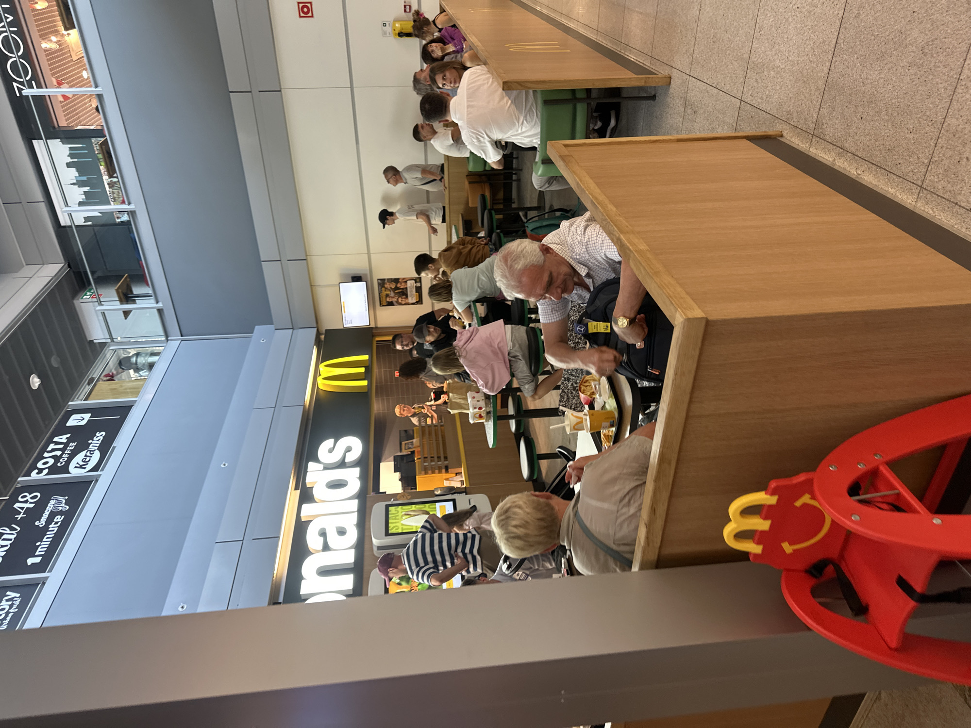The image depicts the interior of a McDonald's restaurant, likely located within an airport given the overhead signage and general architectural style. In the foreground, a red child's high chair with a "Happy Meal" character design is visible. The seating area of the restaurant is populated with numerous patrons. Several individuals are seated at tables, some are in conversation, and others appear to be eating or waiting for their food. The McDonald's branding, including the iconic golden arches and "McDonald's" lettering, is prominently displayed on the restaurant's facade. The environment appears to be a busy commercial space with a mixture of natural and artificial lighting. The overall scene suggests a typical dining experience at a fast-food establishment. Based on the presence of Polish language on some of the overhead signage, the location is confirmed to be Warsaw, Poland.