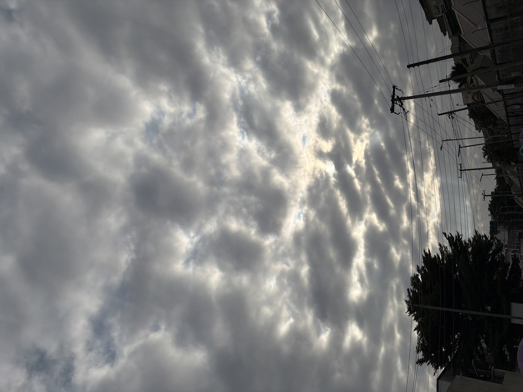 The image depicts a view looking upwards at a cloudy sky over Igboefon, Nigeria. The clouds are dense and gray, with patches of sunlight breaking through in several areas. Below the sky, power lines and utility poles are visible, crisscrossing the frame. A large, dark silhouette of a tree stands prominently on the left side of the image. In the background, the rooftops of buildings and more utility poles can be seen, suggesting an urban or suburban setting. The overall atmosphere is subdued, likely due to the overcast weather.