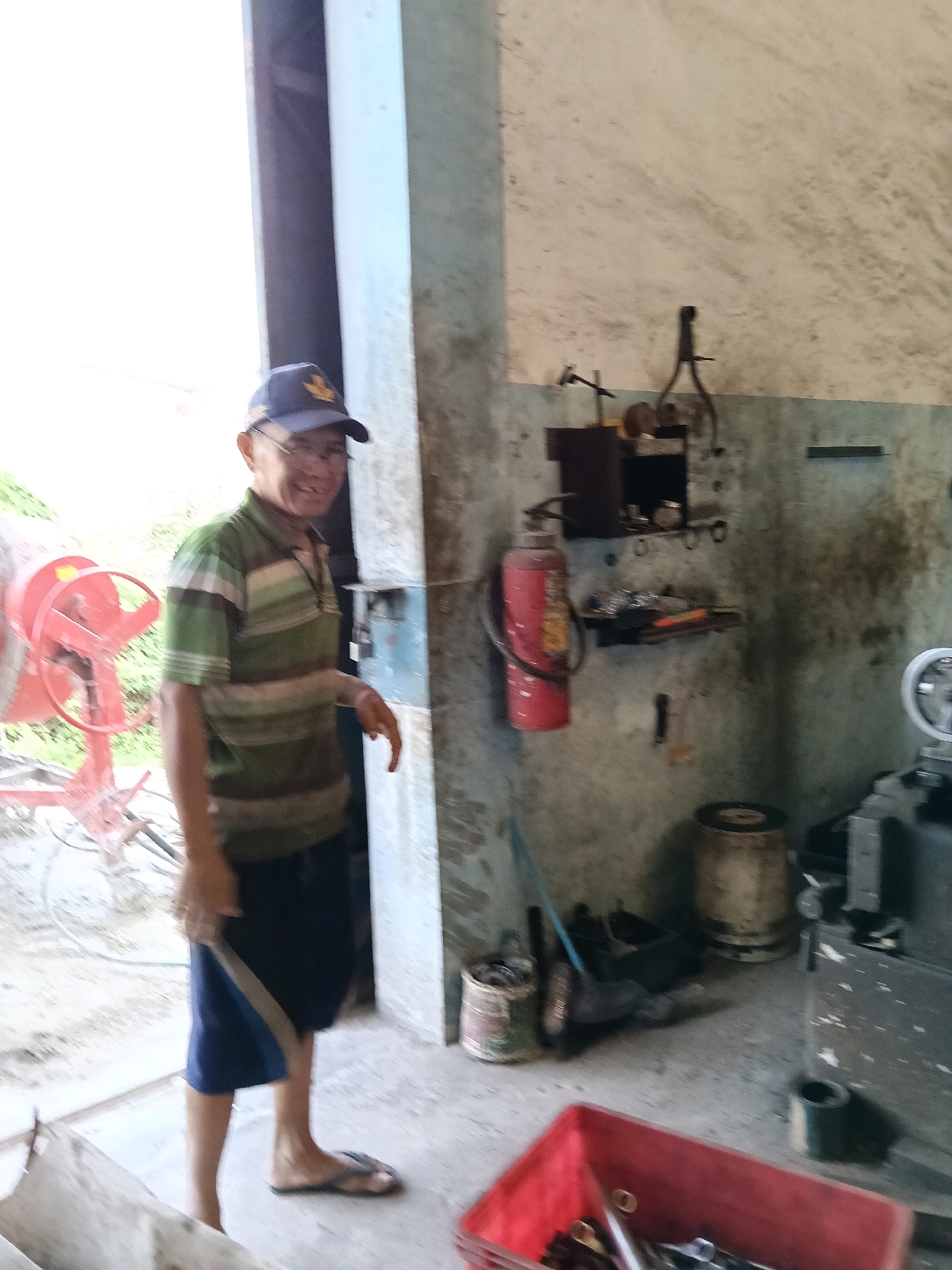 A man wearing a green and white striped polo shirt, blue shorts, a blue baseball cap, and glasses stands in a workshop. He is holding a piece of metal. To his left is a red cement mixer. The workshop walls are concrete and appear weathered. Shelves on the wall hold various tools and equipment, including a red fire extinguisher. In the foreground, a red plastic bin is filled with metal parts. A metal lathe is visible on the right side of the image. The floor is concrete. The scene suggests an industrial or repair setting. The specific location in Belimbing, Indonesia, is not evident from the visual information provided.