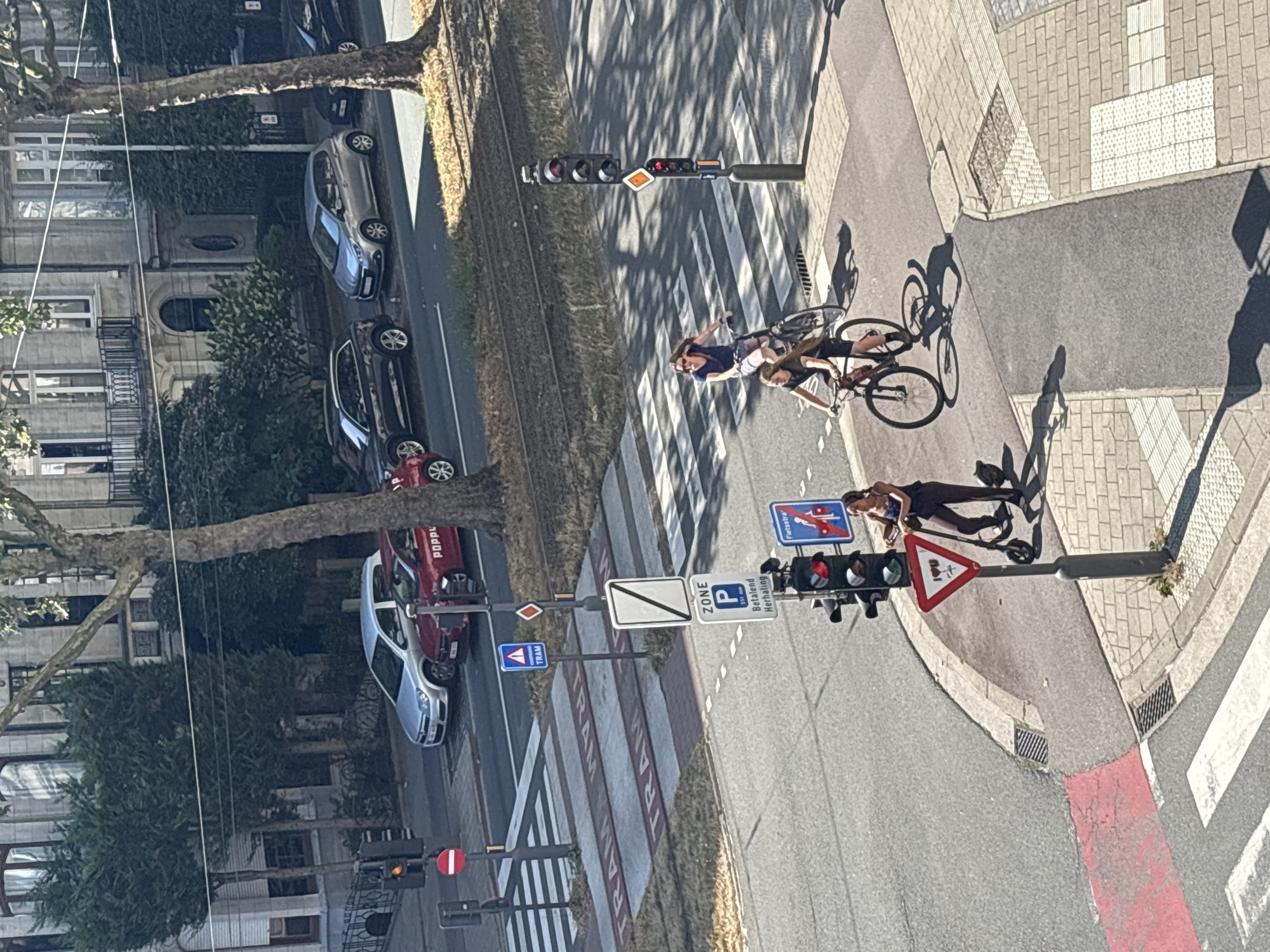 The image captures a sunny day scene at an intersection in Antwerp, Belgium, near the intersection of Harmoniestraat and Tolstraat. Three individuals are present: two women on bicycles and one person on an electric scooter. One woman, wearing a blue dress and sunglasses, is riding a bicycle and appears to be looking to her left. Another woman, dressed in a dark top and shorts, is riding a bicycle behind her. The third individual, wearing a dark top and pants, is on an electric scooter and is looking to their right.
The intersection features tram tracks, pedestrian crossings, and traffic signals. One traffic signal is red, while another appears to be flashing orange. Road signs indicating "ZONE P" (parking zone), "Betalend Herhaling" (paid parking), and "Fietsstraat" (bicycle street) are visible. Several cars are parked along the street, including a red car with the word "POPL..." partially visible on its side, and a silver car. Buildings with balconies and trees line the street, suggesting a residential or commercial area. The scene is viewed from an elevated perspective.
