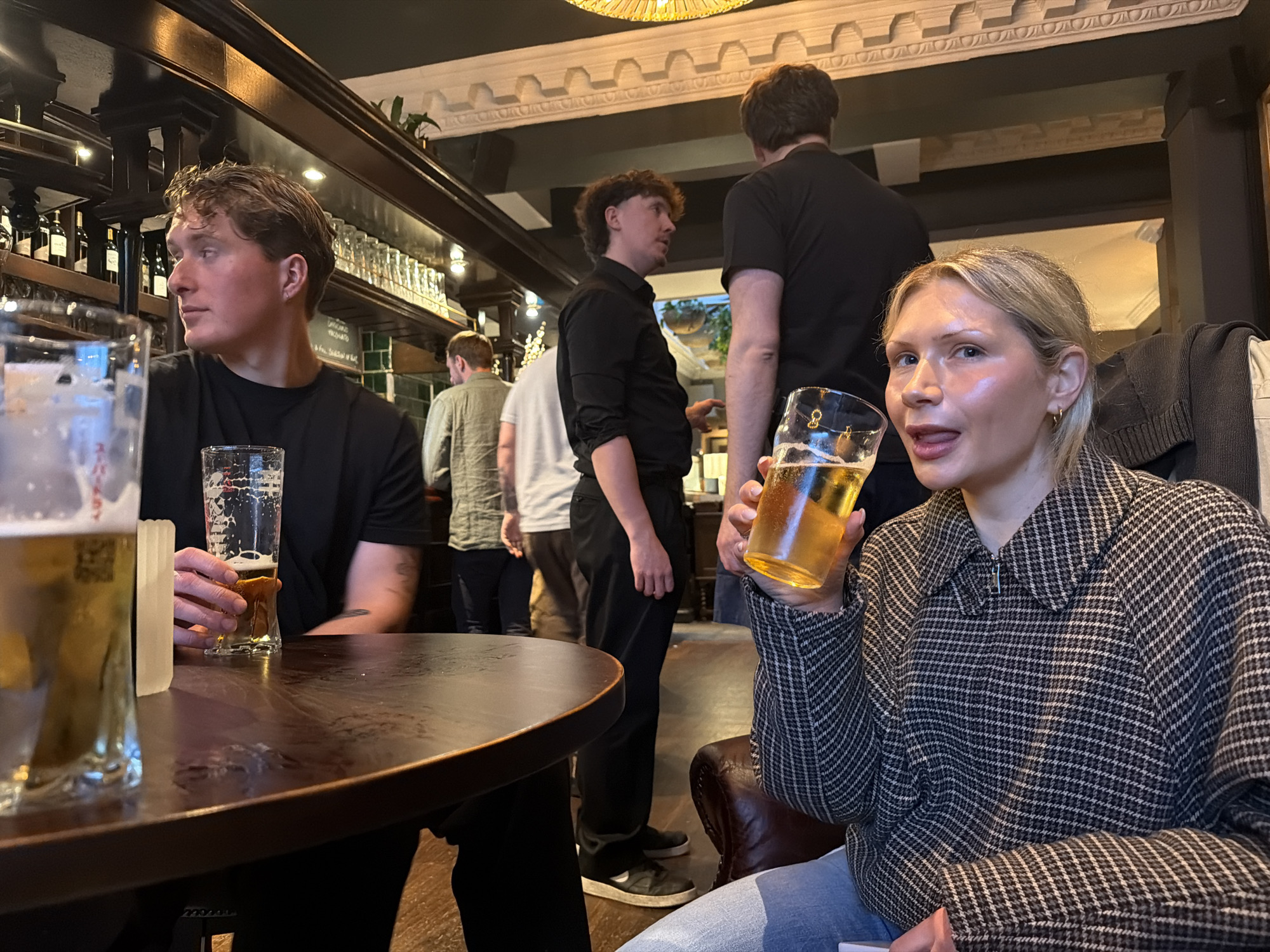 A candid, indoor shot shows a group of people inside what appears to be a pub or bar. A fair-skinned, blond woman is seated on the right, holding a glass of golden liquid, possibly beer. She is wearing a checkered jacket and light-colored jeans, and her tongue is slightly visible. To her left and slightly behind, stands a man in a black shirt, facing away from the camera. Another man, also in a black shirt, is standing further back, facing partially to the side. To the left of the woman, a man with dark curly hair is seated at a round wooden table, holding another beer. In the background, other individuals are visible, with one wearing a light green shirt and another in a white shirt. Behind them, a bar is lined with bottles and glassware. The location is consistent with a pub or bar, possibly in the city of London, United Kingdom.