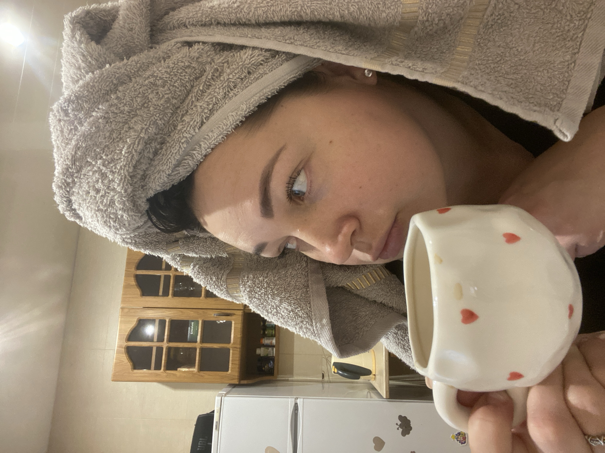A person with a towel wrapped around their head is holding a white mug with red hearts. The background includes a kitchen with wooden cabinets and a white refrigerator decorated with magnets. The location is not discernible as Sochi, Russia from the given image.