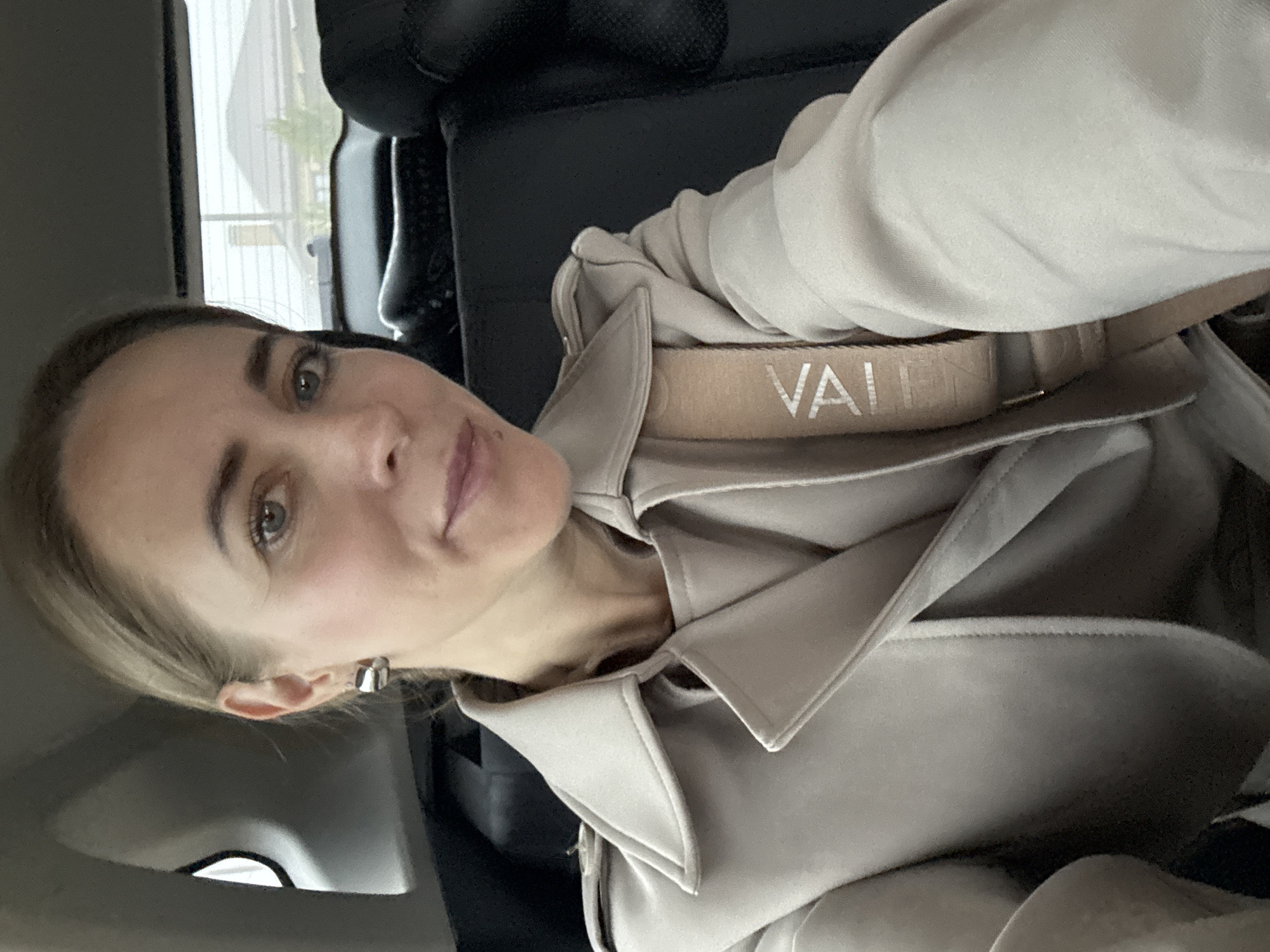 A woman is captured in a close-up, vertically oriented portrait or selfie, seated within the interior of a vehicle, potentially in Chaikovsky, Russia. She has light brown hair, fair skin, and light eyes, looking directly at the camera with a calm, subtle smile. Her right earlobe shows a silver-toned, C-shaped earring.
She is wearing a light beige or tan-colored trench coat or similar outerwear, with its collar and folded fabric visible. Across her chest and shoulder, a matching beige strap is prominently displayed, featuring the word "VALENTINO" in silver or white lettering.
The background consists of dark car upholstery, likely a seat or headrest. Through a window to the left, a blurred glimpse of outdoor scenery suggests buildings or houses under an overcast daylight sky. The image focuses on the woman and her attire, with the car interior serving as the immediate setting.