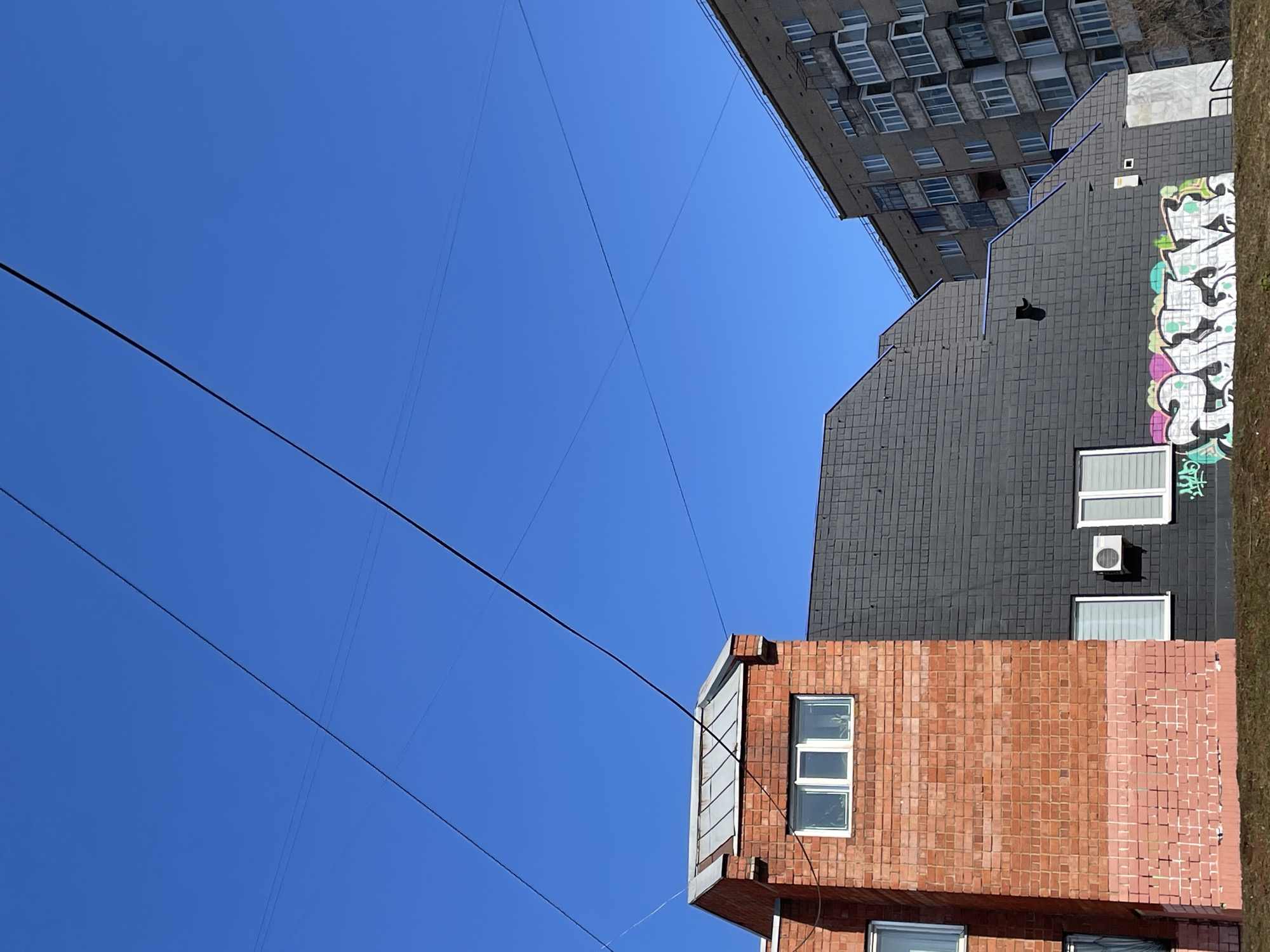 The image depicts a low-angle view of urban architecture against a clear blue sky. Three buildings are visible: a brick building, a grey brick building, and a building with a concrete facade with many windows. The buildings are aligned vertically in the image. Graffiti is present on the grey brick building, and power lines run diagonally across the sky. Based on architectural style and graffiti, the location is likely Izhevsk, Russia.
