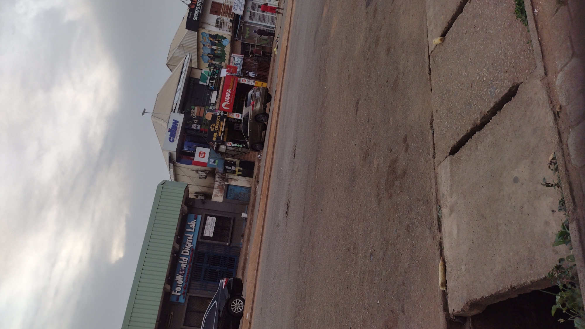 The image depicts a street scene, likely in a commercial area, in Jos, Nigeria. On the left side, there is a "FotoWorld Digital Lab" with its gate closed. Further up the street, there are various businesses including "CELEN", "MMB CONNECT", and "SPORTS STORE", all with advertisements. A few cars are parked along the roadside. The road appears to be paved, with cracks visible on the sidewalk. The sky is overcast. One person is walking on the sidewalk near the stores.