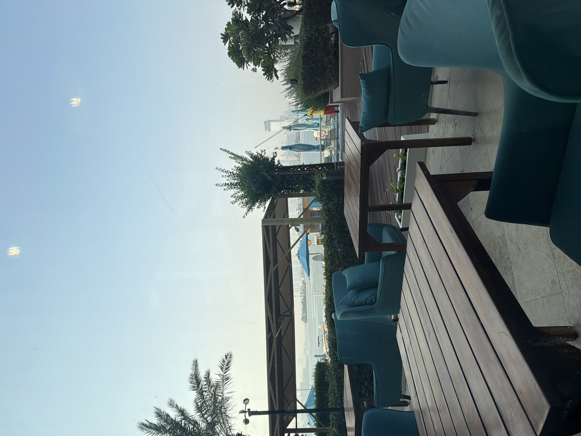 The image depicts an outdoor seating area with a view of the Dubai skyline across a body of water. In the foreground, there are several dark wooden tables and teal upholstered chairs with pillows. The tables have a slatted surface. Beyond the seating area, there is a line of lush green bushes and trees. In the middle ground, several large blue beach umbrellas are visible, along with lounge chairs and a swimming pool. A person in a yellow shirt and red pants is cleaning or tending to something near the pool area, holding a long pole. The background features a hazy view of the Dubai cityscape, characterized by tall buildings. Palm fronds from a tree on the left side of the frame extend into the upper left corner. The sky is a pale, hazy blue. The overall scene suggests a resort or hotel setting, possibly overlooking a marina or beach, within Dubai, United Arab Emirates.