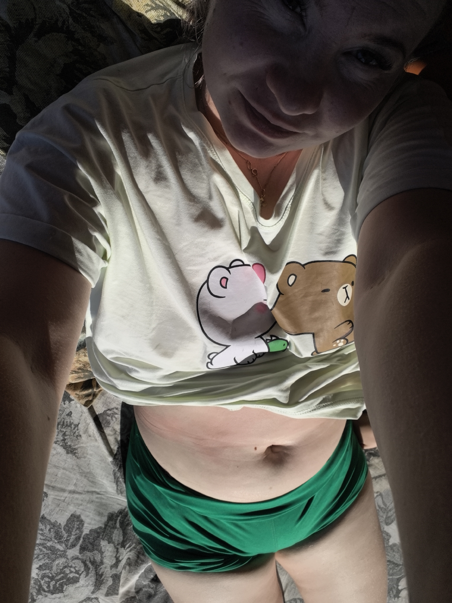 A woman is lying down, taking a selfie. She is wearing a light green t-shirt with cartoon bears printed on it and green shorts. Her midsection and face are visible, along with her arms and legs. The background appears to be a bed with a patterned blanket. The lighting suggests she is indoors, possibly near a window due to the shadows cast on her and the shirt. There is no indication of the city of Vityazevo, Russia in the image.