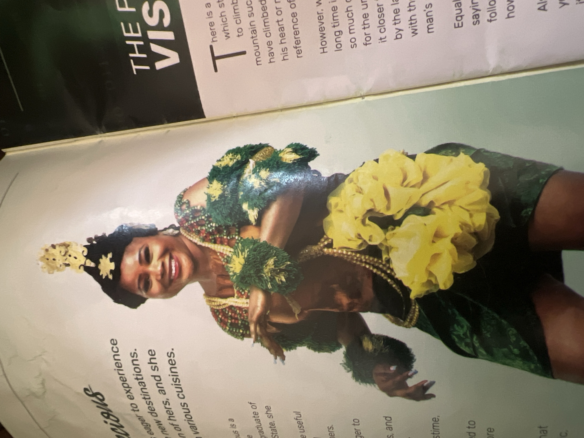 The image shows a woman in traditional attire, likely for a cultural event or performance, against a neutral background with text on either side. The woman is wearing a dark headdress adorned with gold-colored ornaments, a multi-colored beaded necklace, and matching beaded bracelets. She also wears green, fluffy armbands with yellow accents, and a green skirt with a leaf-like pattern. A large, yellow, ruffled accessory is attached to her right side. Her skin tone is dark, and she has a bright smile.
The text on the left side is mostly legible and appears to be descriptive, mentioning eagerness to experience new destinations and various cuisines. The text on the right side features large, bold font spelling "THE P VIS," suggesting a headline or title. Below this, there is smaller text that begins with "T here is a" and continues with descriptive sentences.
There is no indication within the image that it is located in the city of Jalingo, Nigeria. The background and surrounding text do not provide any geographical context.
