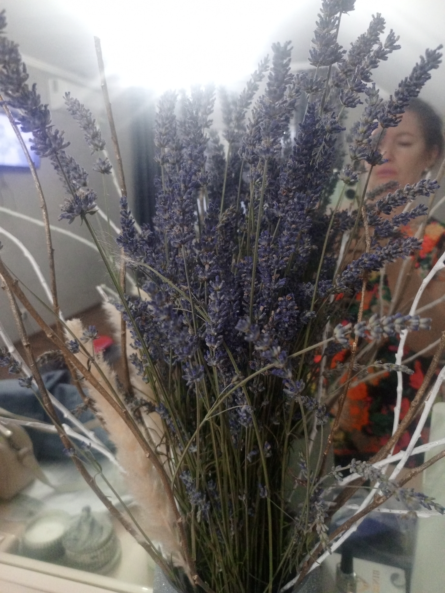 A close-up, eye-level, indoor shot shows a bouquet of dried lavender and decorative branches in a vase. The lavender stalks are thin and upright, with clusters of small purple flowers. Some of the branches are bare and brown, while others are painted white. In the background, a woman with dark hair is out of focus, wearing a colorful, patterned shirt. The scene appears to be set in a room, possibly a home, in Sovkhozny, Russia, as indicated by the context. The lighting is bright, with a glare on the left side of the image, suggesting a light source is present.