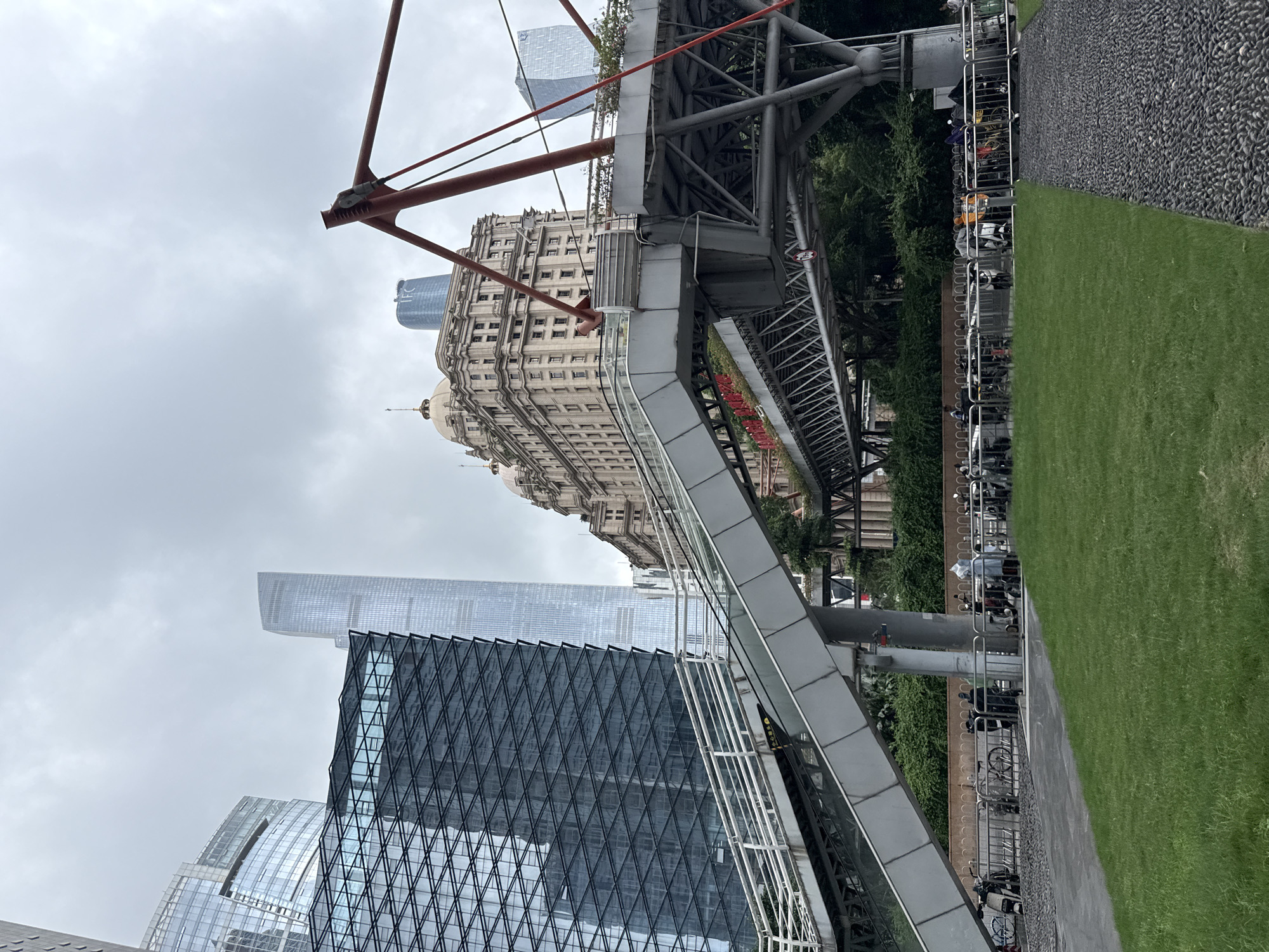The image depicts an urban scene in Tianhe District, China, characterized by a mix of modern and historical architecture under an overcast sky. A prominent feature is a large, elevated walkway or bridge structure made of metal, with red support beams. This structure appears to be a pedestrian walkway, possibly connecting different parts of the urban landscape or an elevated park.
Below the walkway, several buildings are visible. A grand, older building with a dome-like structure and intricate stonework stands out, contrasting with the sleek, glass-fronted skyscrapers surrounding it. One skyscraper has a distinctive diamond-patterned facade. Another tall, modern building in the background appears partially obscured by the haze.
The foreground shows a neatly manicured green lawn bordered by a dark, pebbled pathway. Adjacent to the lawn and pathway is a long, silver fence, behind which numerous bicycles are parked, suggesting a public area or a bike-sharing station. A few individuals can be seen near the bicycles. The overall atmosphere is subdued, likely due to the cloudy weather, which obscures the time of day and lends a somewhat muted tone to the scene. The presence of contemporary urban infrastructure alongside historical buildings suggests a city that is both forward-looking and mindful of its past.