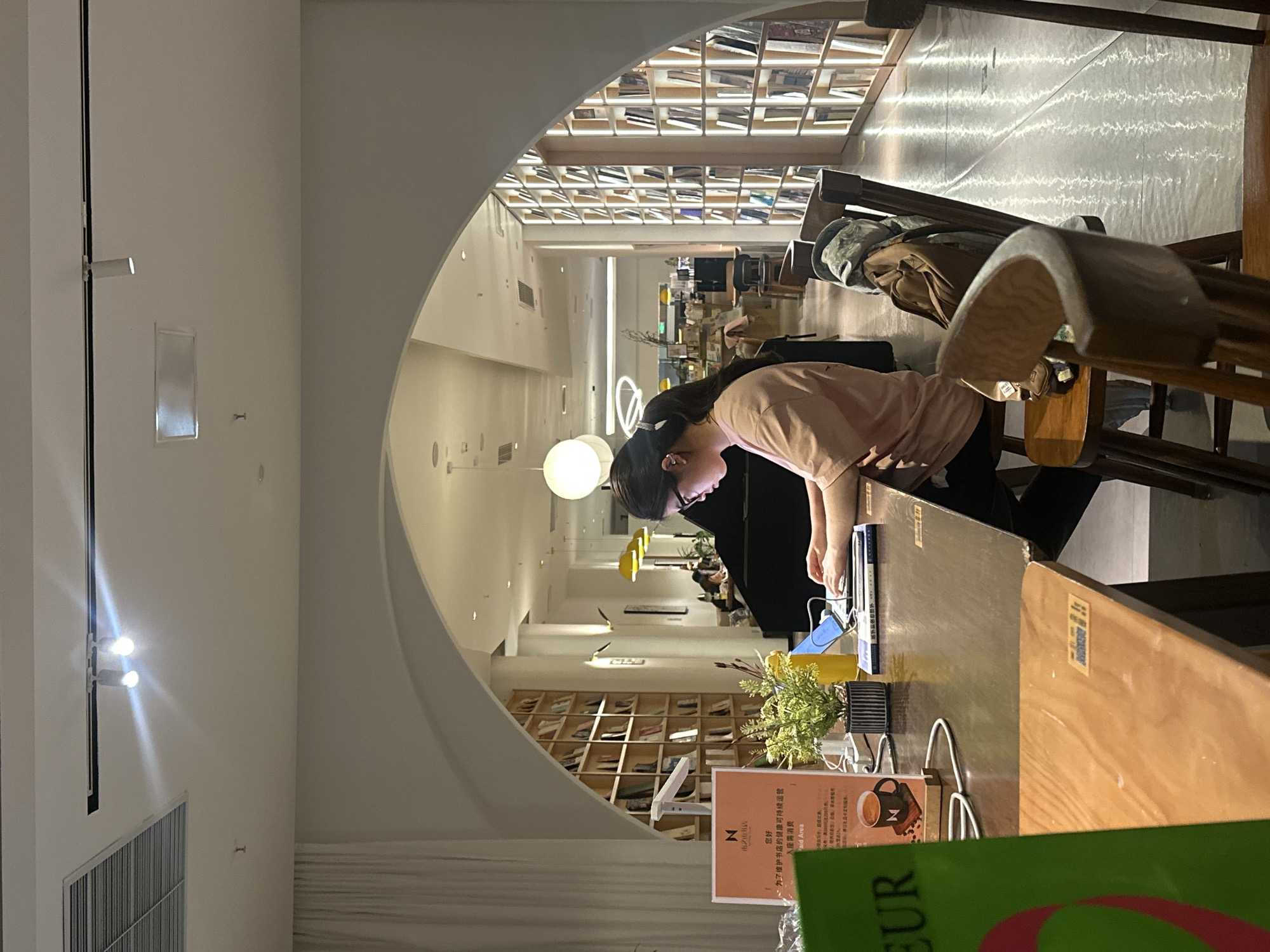The image depicts an interior scene of what appears to be a library or cafe, possibly located in the Chongqing High-tech Zone, China. In the foreground, a woman with dark hair pulled back into a ponytail is seated at a dark wooden table, facing a laptop. She is wearing glasses and a light pink t-shirt. Her hands are positioned on the keyboard, suggesting she is actively working or studying.
The table is cluttered with various items, including the laptop, a blue book, a plant in a striped pot, a yellow bottle, and a sign with Chinese characters and a logo featuring the letter "N" inside a coffee cup. A green item, possibly a book or menu, is partially visible in the lower left corner of the frame. The background reveals shelves filled with books, characteristic of a library or bookstore. Archways divide the space, with warm lighting emanating from ceiling fixtures. The overall atmosphere is one of quiet productivity or leisure.