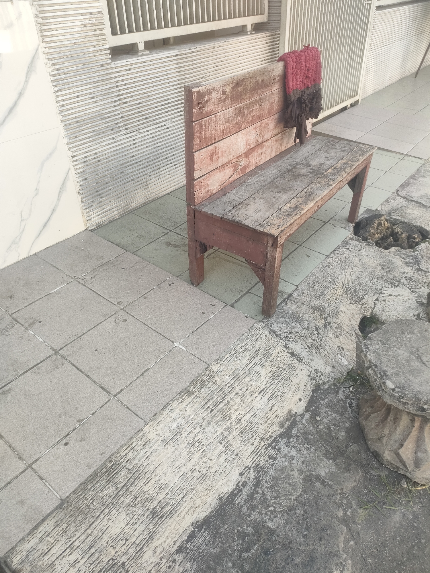 A wooden bench with a reddish-brown frame and weathered planks is positioned on a tiled patio in Surabaya, Indonesia. A red and brown patterned cloth is draped over the backrest of the bench. To the right of the bench, a cat is sleeping on the ground. The patio area features a mix of tiled and concrete surfaces, with some rough textures and cracks visible in the concrete. In the background, a white metal gate and a textured wall are present. The overall scene suggests an outdoor space, possibly a residential or public area in the city.