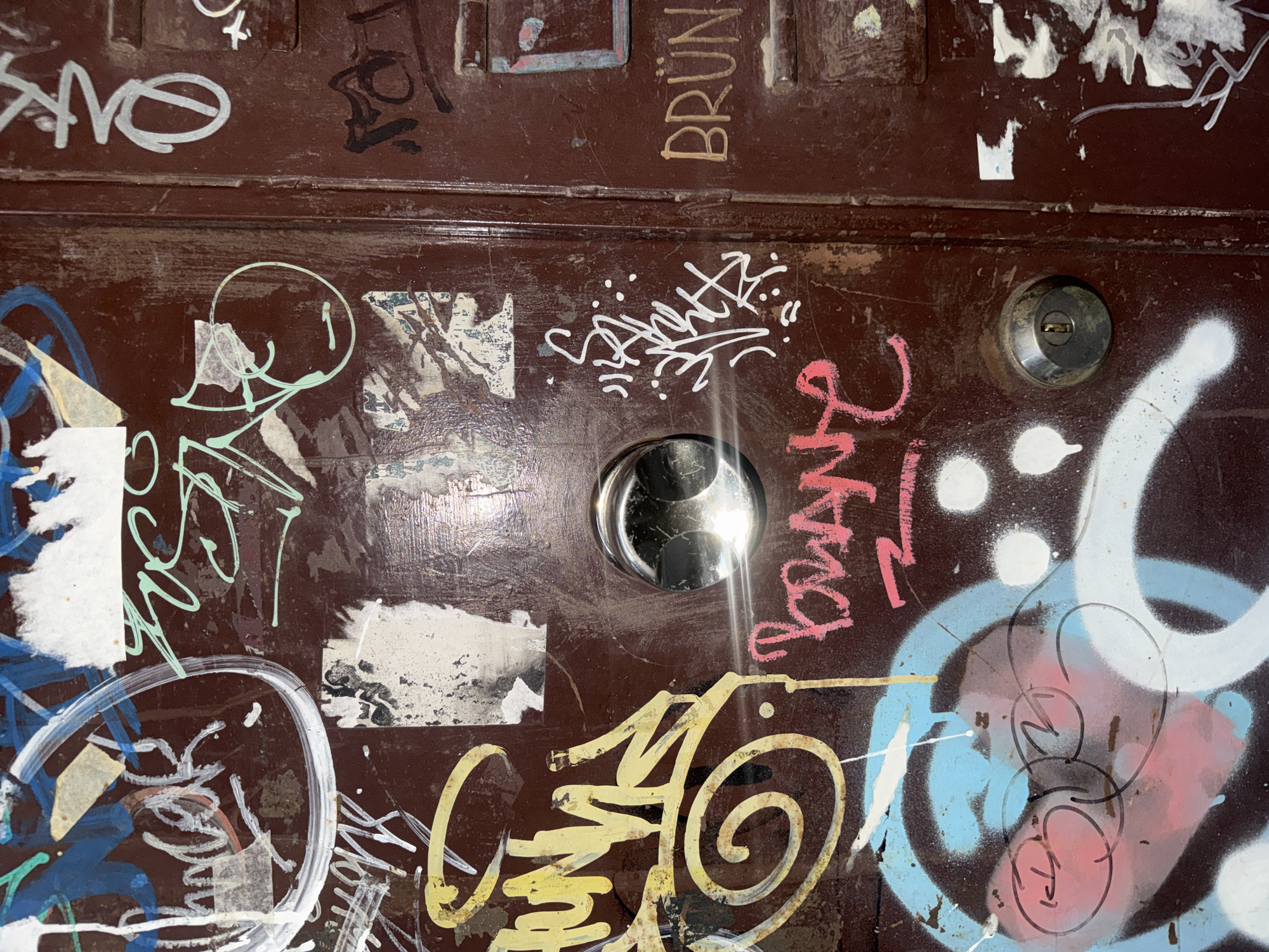 A close-up shows a brown door heavily covered in graffiti, with a peephole and a keyhole lock. The graffiti is multicolored, featuring tags in white, yellow, blue, and pink. Some stickers or paper remnants are also visible. The door appears to be weathered and possibly located in an urban area, indicated by the accumulation of street art. The word "BRUN" is visible in the top right corner. No people are visible in the image. The location is Sofia, Bulgaria.