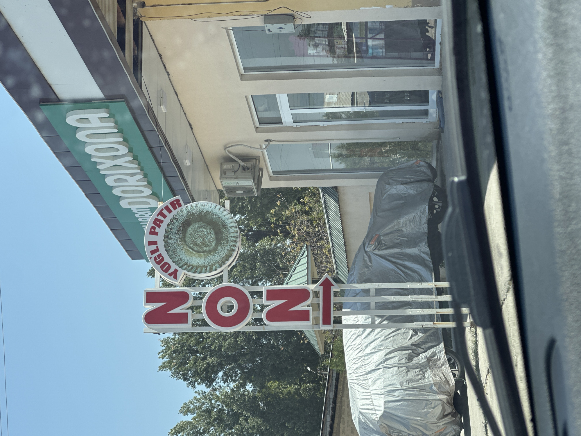 The image captures a street scene in Tashkent, Uzbekistan, on a clear day. In the foreground, the windshield and wipers of a vehicle are visible, indicating the photo was taken from inside a car. Beyond the car, a building with a yellow facade and large glass windows is present. Above the building, a green sign with white, raised lettering reads "DORIXONA," which translates to "pharmacy." To the left of the building, a vertical sign with red letters spells out "ZON." A circular sign with a patterned design is positioned next to the "ZON" sign, and a red arrow points to the right. Two cars are parked in front of the building, both covered with grey tarpaulins. Trees line the background, and part of a green roof is visible behind the covered vehicles. The overall impression is that of a commercial area, likely a street in Tashkent.
