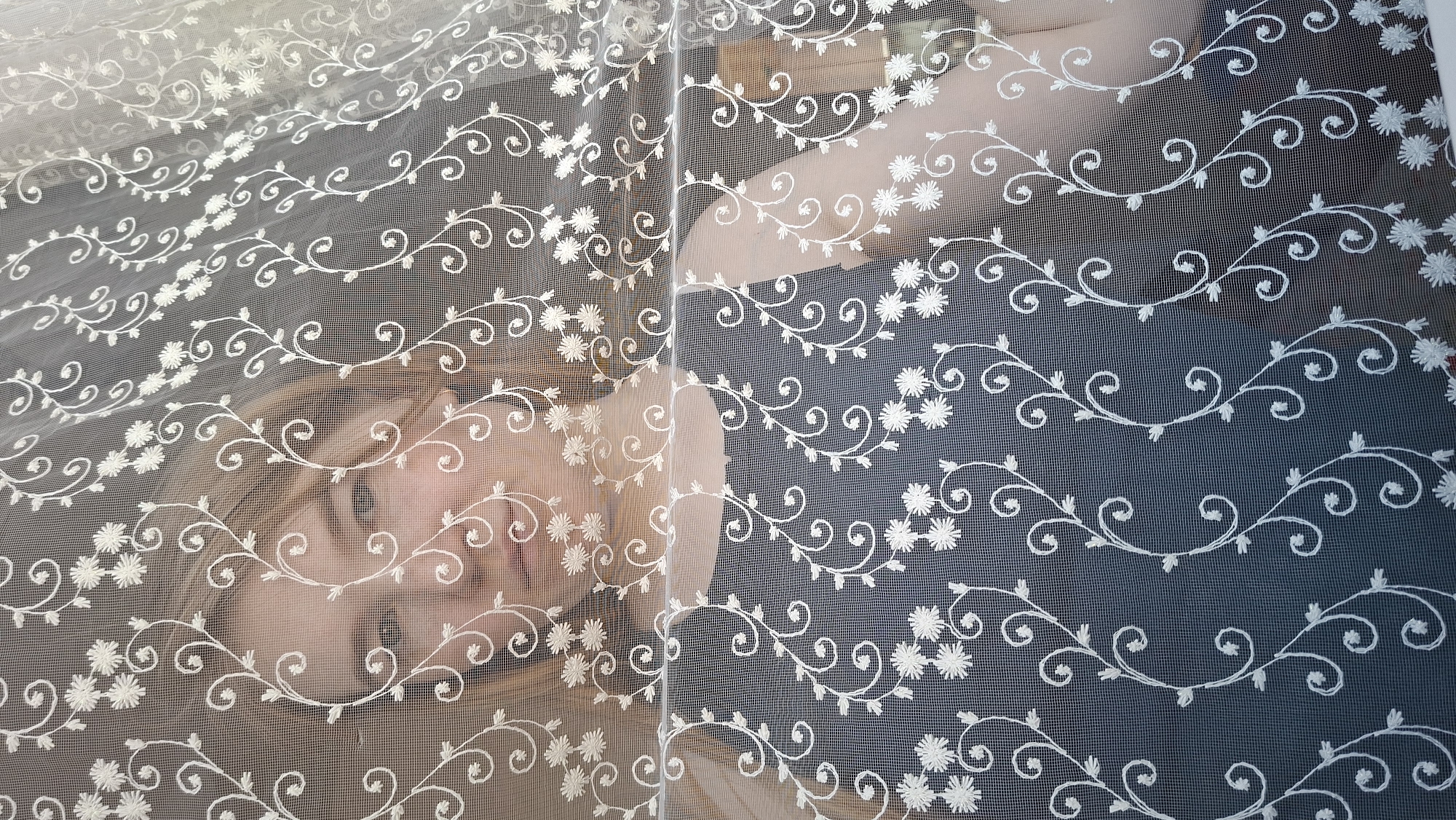 A person with light skin and light brown hair is visible behind a sheer white curtain with an embroidered floral and vine pattern. The person's face, particularly the nose and mouth, is somewhat obscured by the curtain. The background behind the curtain appears dark, possibly a wall or other surface.
The location of the image is Большой Букор, Russia.