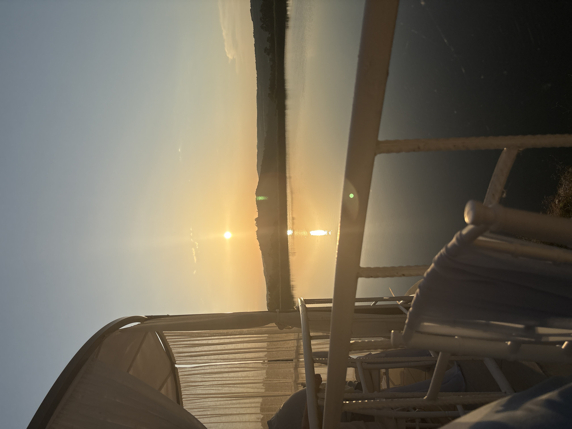 The photograph captures a serene sunset over a body of water, viewed from a covered balcony or patio. The sun, a bright orb, is low on the horizon, casting a warm, golden glow across the sky and reflecting on the calm surface of the lake. Rolling hills, densely covered in trees, form a natural backdrop to the water.
In the foreground, a white railing runs horizontally across the frame, separating the viewing area from the water. To the left, the edge of a white, curtained structure, possibly a gazebo or cabana, is visible, its light fabric gently swaying. Several white chairs are arranged on the patio, some draped with light blue fabric. The overall atmosphere is tranquil and peaceful, suggesting a moment of quiet contemplation or relaxation. The scene is located in Skalsko, Bulgaria.