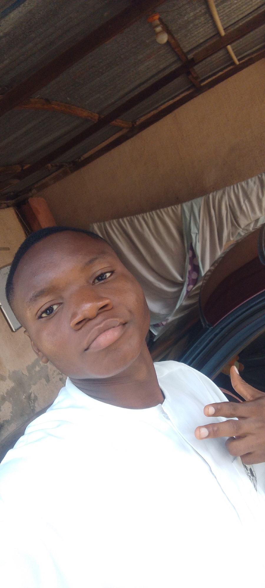 A young Black male is pictured in a close-up, selfie-style shot, looking directly at the camera. He is wearing a white collared shirt. His left hand is raised, with two fingers extended in a peace sign. The background suggests an outdoor or semi-outdoor setting with a corrugated metal roof supported by wooden beams. Draped fabric, possibly a curtain or sheet, hangs in the middle ground, obscuring a dark vehicle behind him. The overall scene appears to be in a humble, perhaps rural or informal, urban environment.