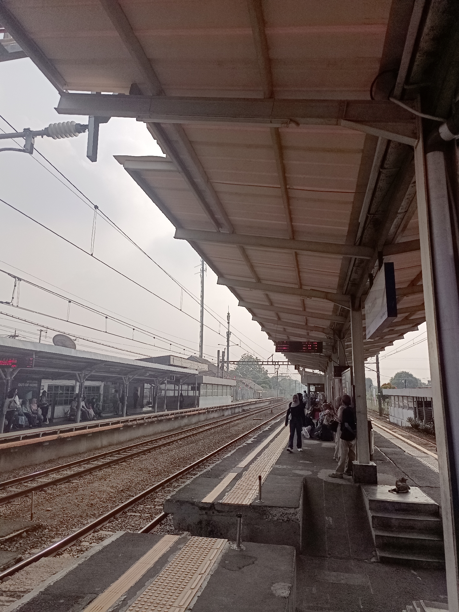 The image depicts a view of a train station platform in South Tangerang, Indonesia, on an overcast day. The platform is covered by a light-colored, corrugated metal roof supported by a steel structure and pillars. Overhead power lines for the train are visible against the hazy sky, suspended from utility poles.
On the platform, several people are waiting. Some are seated on benches under the covered area, while others are standing. The individuals are dressed in casual attire. In the foreground, the train tracks run parallel to the platform. The tracks are laid on a ballast of crushed stone. Tactile paving, designed for visually impaired individuals, is present along the edge of the platform.
Across the tracks, another platform and a building with windows are visible. To the right of the platform, a staircase leads down from the elevated platform. A small, dark object, possibly a cat, is resting on one of the steps. The overall scene suggests a functional public transportation hub.