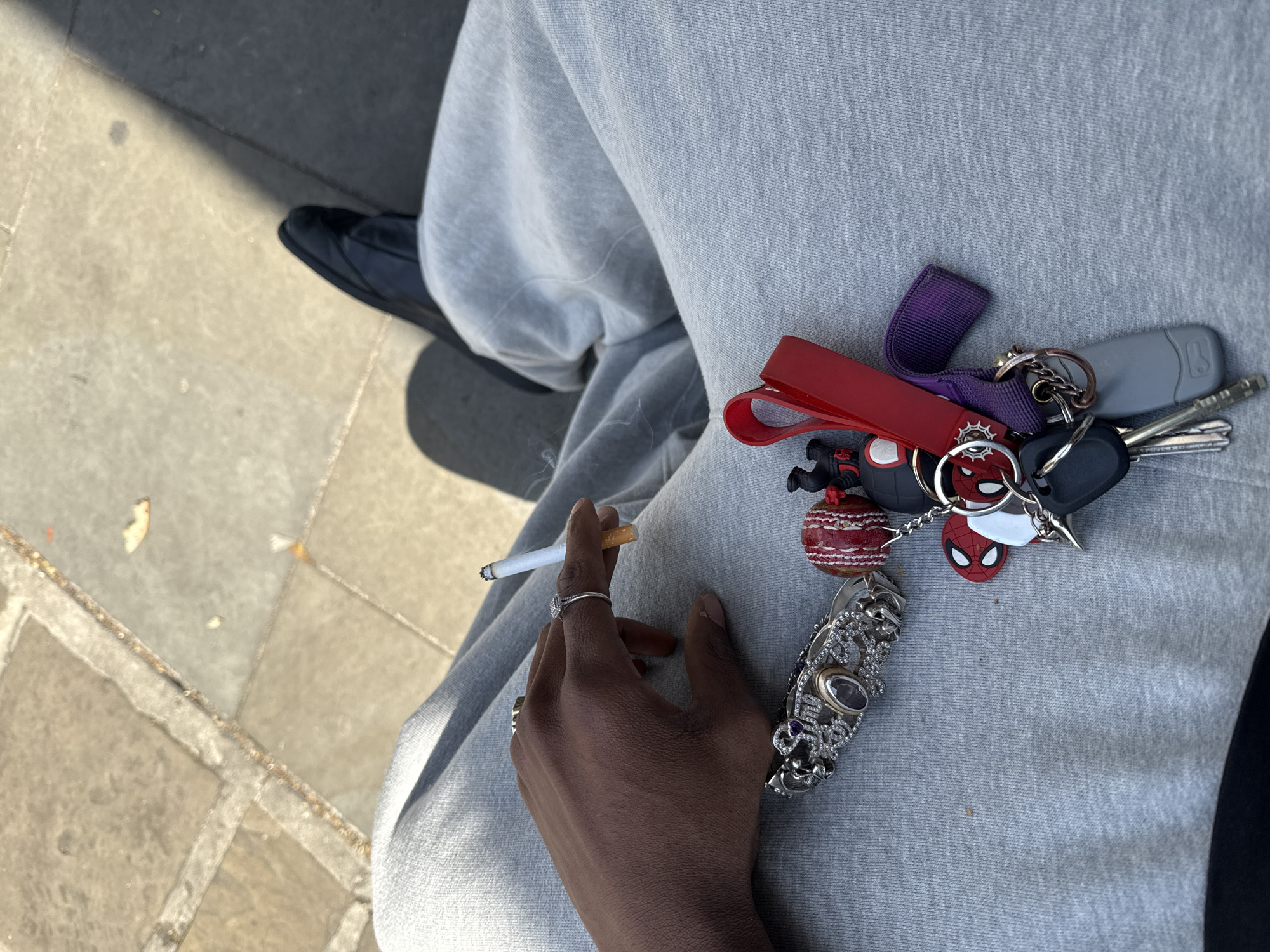 The image presents a high-angle, close-up view of a person's hand, leg, and a collection of keys in an outdoor setting. The subject, a person with dark skin, is visible from the upper leg down. They are wearing light grey sweatpants and a dark shoe is partially visible on the ground. A silver ring is worn on the person's right index finger.
The primary activity is the person holding a lit cigarette between their index and middle fingers, suggesting they are smoking or preparing to smoke. Resting on their lap, on the grey sweatpants, is a large bunch of keys attached to a metal ring. This collection includes various keychains: a vibrant red strap, a purple fabric loop, a grey car key fob, multiple standard metal keys, a small red and black Spider-Man head, a spherical red and white charm resembling a cricket ball, and an ornate, detailed silver key ornament with purple gemstones.
The scene is brightly lit by natural sunlight, casting distinct shadows on the paved ground, indicating it is daytime and sunny. The ground is made up of light-colored, square paving slabs with visible grout lines, common in urban or public outdoor areas. No discernible text is visible within the frame, and the person's emotions cannot be determined from this partial view. The context places this scene in London, United Kingdom.