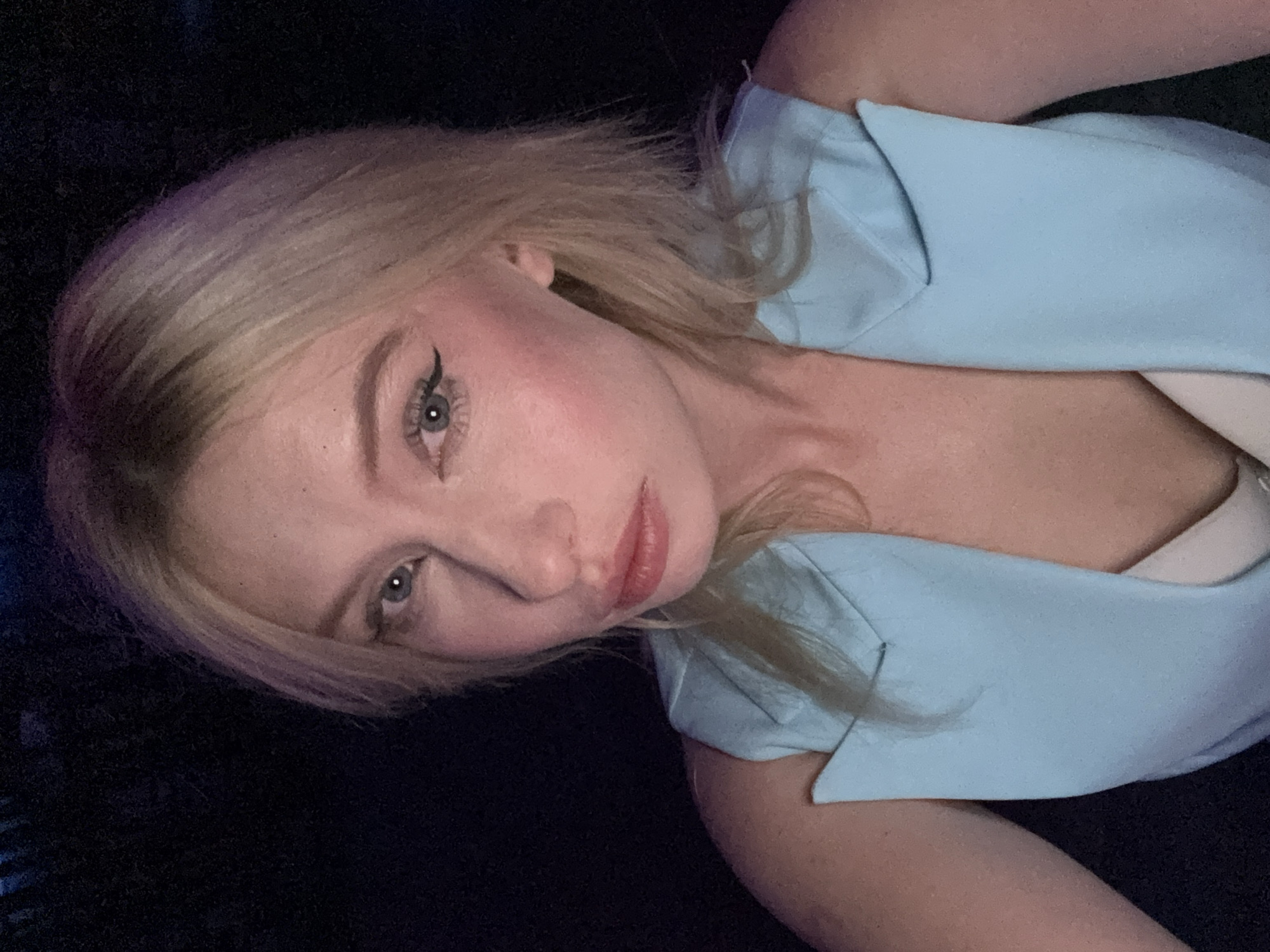 A close-up selfie of a young woman with blonde hair, blue eyes, and fair skin, taken against a dark background. She is wearing a light blue sleeveless jacket with a white garment underneath. Her makeup includes winged eyeliner and blush. The lighting is dim, with some purple tones visible in her hair. The scene is not identifiable as being in Amman, Jordan.