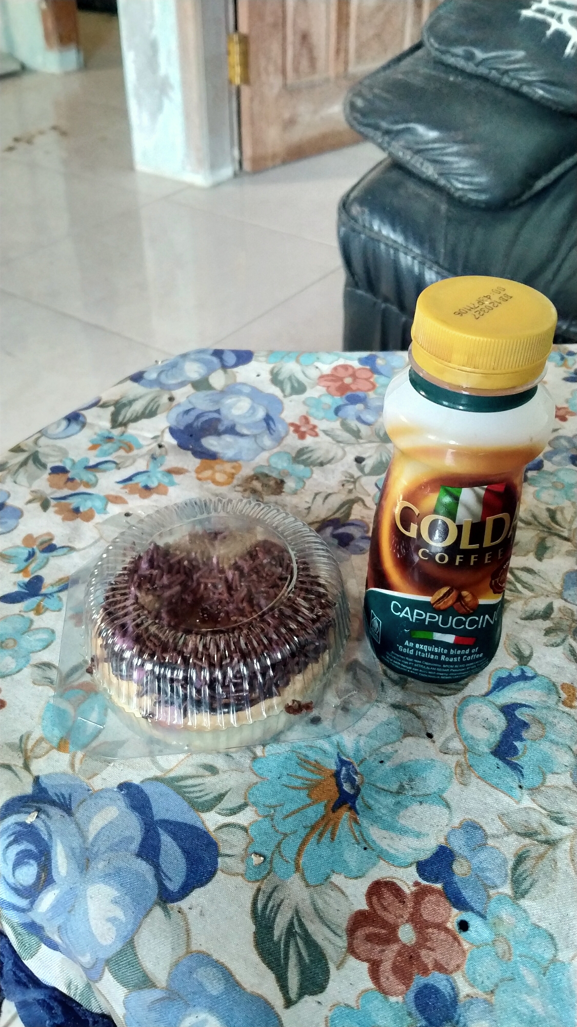 A bottle of Gold Coffee Cappuccino and a small cake are placed on a floral-patterned tablecloth. The bottle is made of plastic and has a yellow cap. The cake is round and covered in chocolate sprinkles. In the background, a black leather couch and a wooden door are visible. The scene appears to be indoors, likely in a residential setting. The location is identified as Wonosegoro, Indonesia.