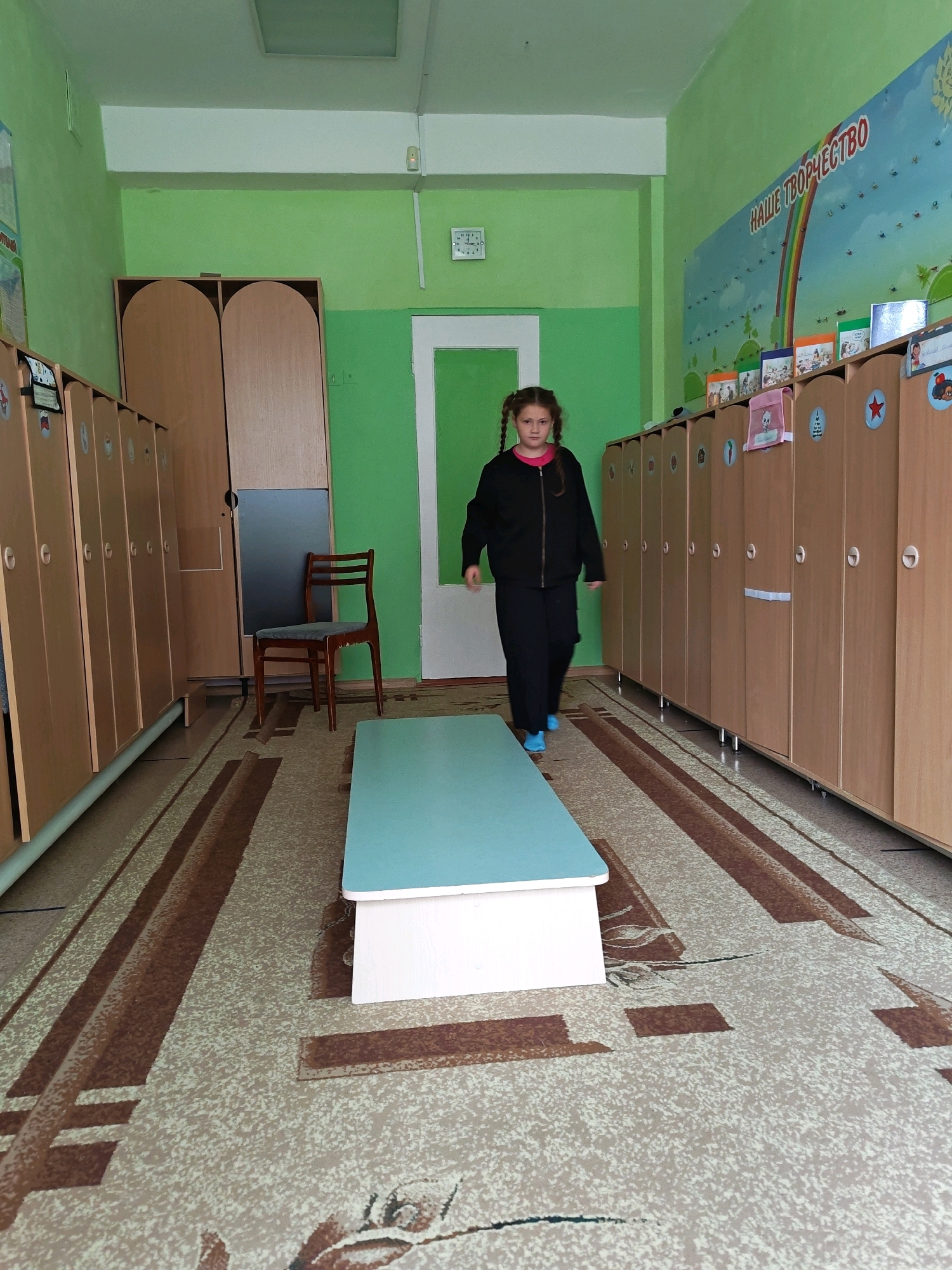 A young girl with braided hair walks through a room that appears to be a school locker room or a changing room. She is wearing a black tracksuit and blue shoes. The room has green walls and a patterned carpet. On one side of the room are rows of wooden lockers, and on the other side, there are more lockers and a large wooden wardrobe. A wooden chair is visible in the background, along with a white door. There is a long, light blue bench in the center of the room. A colorful mural of a rainbow and text is on the far wall. A clock is mounted on the wall. The overall impression is of an institutional or educational setting. The location of Chaikovsky, Russia is not explicitly indicated within the image itself and is provided as external context.