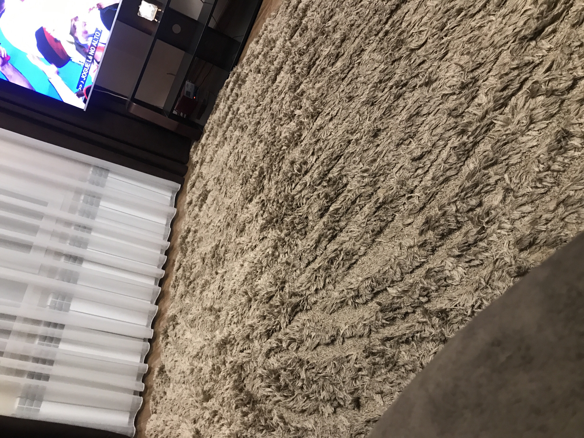 The image shows a section of a room with a large, shaggy, light brown rug covering much of the visible floor space. Adjacent to the rug are partially visible floor-to-ceiling white curtains obscuring windows. A black entertainment center, containing a television screen showing figures in motion, is positioned near the curtains. The angle of the shot suggests the camera is low, possibly near floor level. Details such as brand names or specific identification of the people on the screen are not discernible. No specific indicators suggest the location is Yerevan, Armenia.