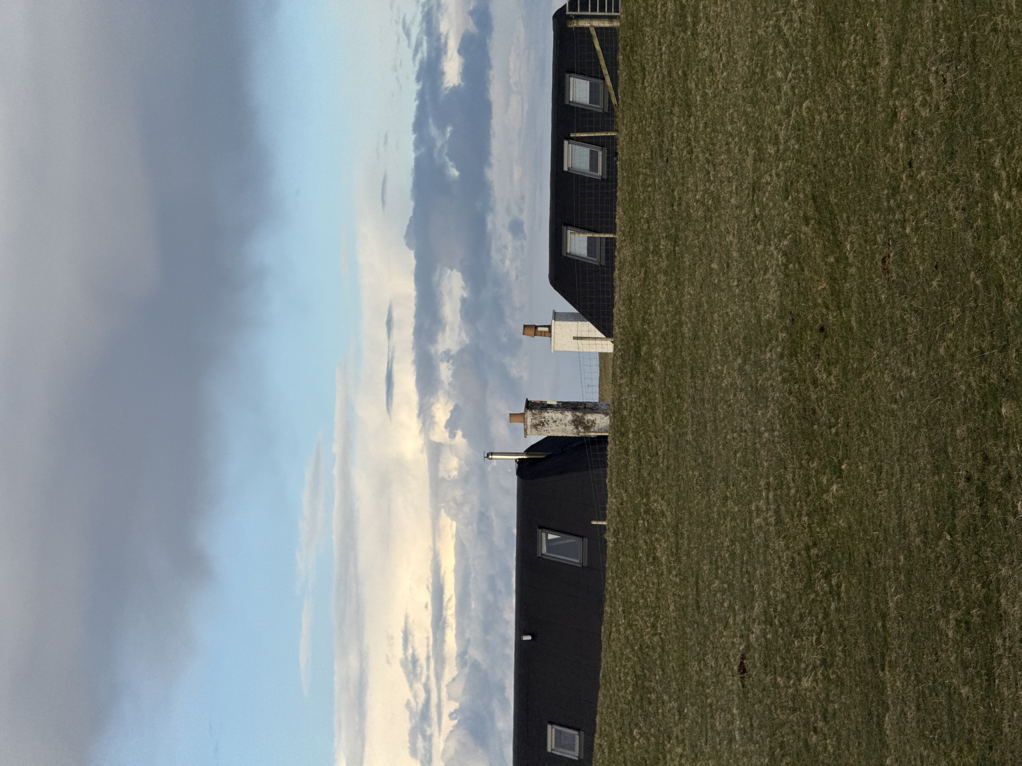 The image shows a rural scene with a field of dry grass on the right and a cloudy sky on the left. There are two buildings visible, both with dark exteriors and skylight windows. One building has a white chimney with a brown top, and the other has a metal chimney. The location is Unknown, Unknown.