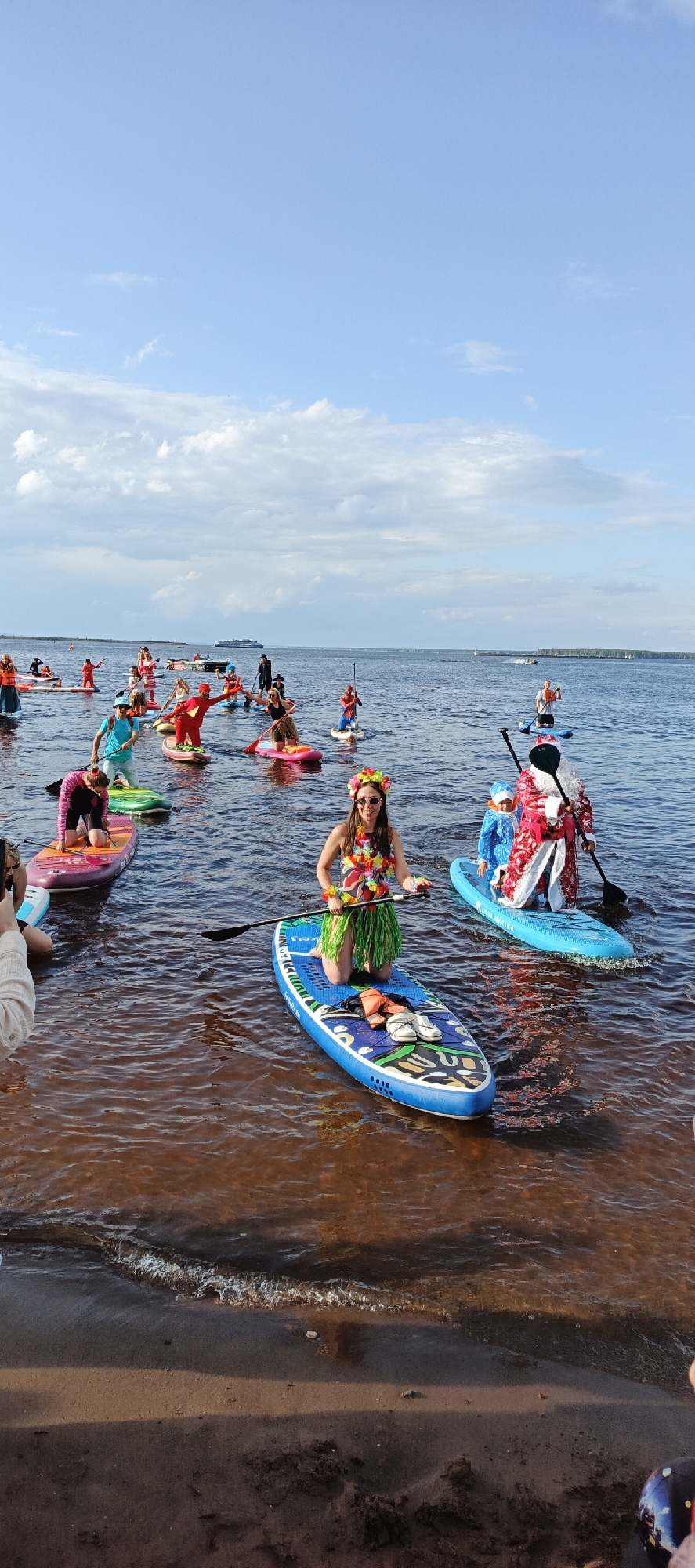A group of people are paddleboarding on a body of water, likely a river or lake. The sky is partly cloudy with a light blue hue, suggesting it is daytime, possibly late afternoon given the warm tones in the water.
Many of the paddleboarders are dressed in costumes. In the foreground, a woman is kneeling on a blue paddleboard, wearing a floral lei around her neck and a grass skirt, giving her a hula dancer appearance. Another paddleboarder further back is dressed as Santa Claus, accompanied by a child in a blue costume. Other individuals are wearing superhero costumes, including one in red and yellow, resembling The Flash, and another in a red and blue Spider-Man costume. Some participants are wearing life vests.
The water appears calm, with ripples reflecting the sunlight and the sky. The shoreline is visible in the lower part of the image, featuring sand and some darker patches, possibly wet sand or soil. In the background, other paddleboarders are spread out on the water, and a larger vessel, possibly a ferry or ship, is visible in the distance on the horizon. The scene evokes a sense of fun and celebration, likely a themed event or festival. No visible text is present in the image.