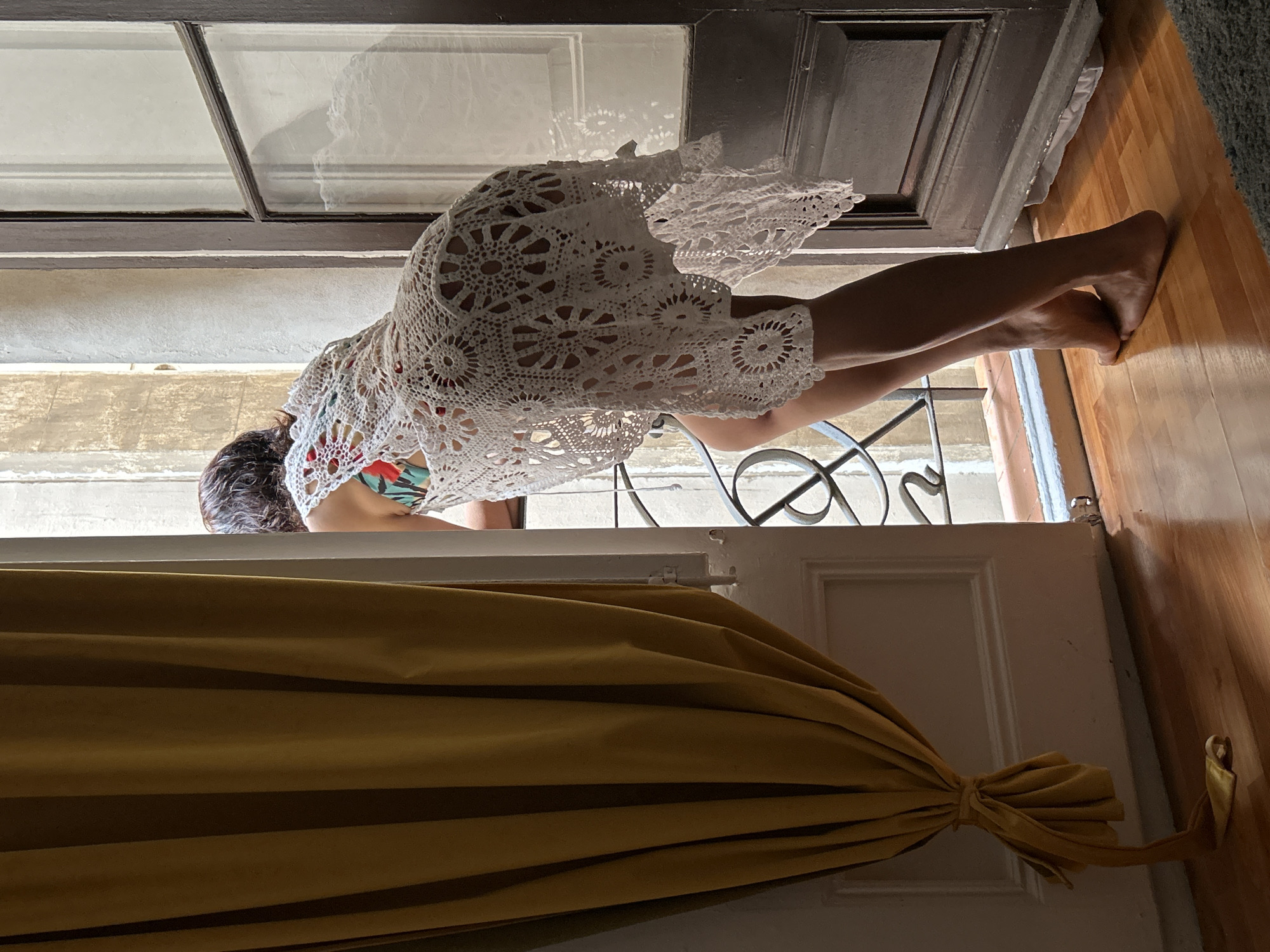 A person wearing a white crochet dress and a colorful bathing suit is leaning out of a window overlooking an external wall. The window has black iron bars. The person is leaning over the window sill. The window is located in Barcelona, Spain. The foreground is dominated by a yellow curtain tied in a knot. The floor inside the room is wooden.