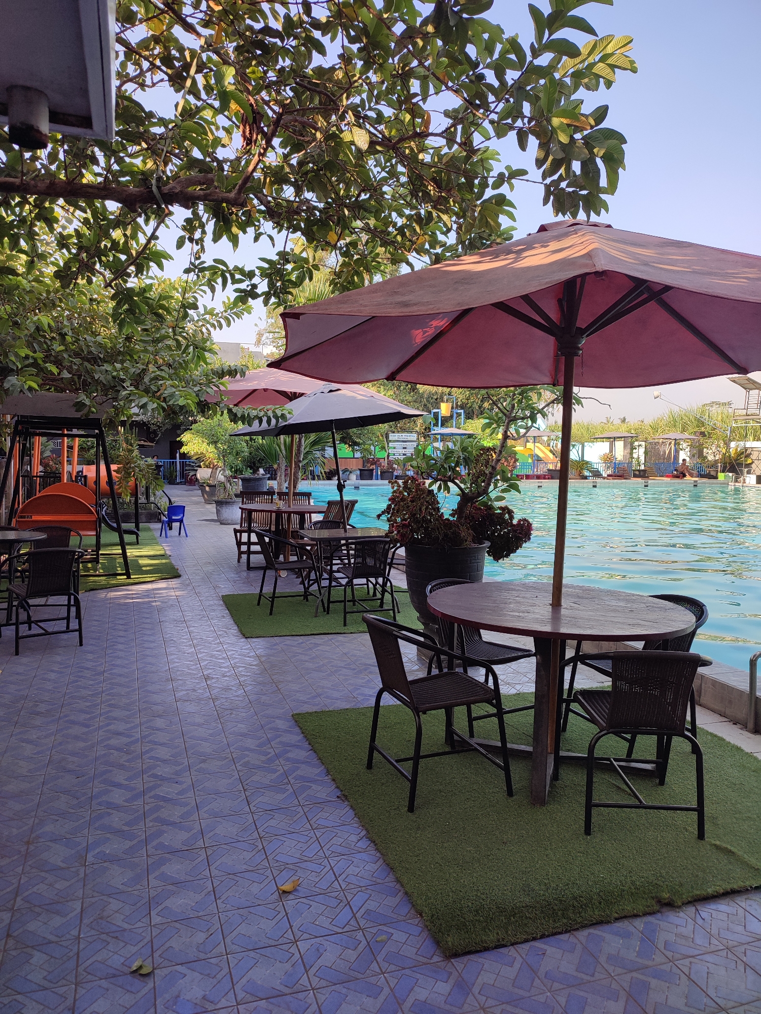 The image depicts an outdoor seating area adjacent to a swimming pool at a resort, possibly in Karangduren, Indonesia. In the foreground, several round tables with chairs are set up on artificial turf patches, bordered by blue tiled flooring. Two large, maroon patio umbrellas provide shade over some of the tables. Further back, past more tables and chairs, is a playground with orange slides and a swing set. Beyond the playground, a large swimming pool with clear blue water stretches across the middle ground, with several resort buildings and palm trees visible in the background. The sky is clear and blue. The overall impression is a leisurely, sunny day at a family-friendly resort.