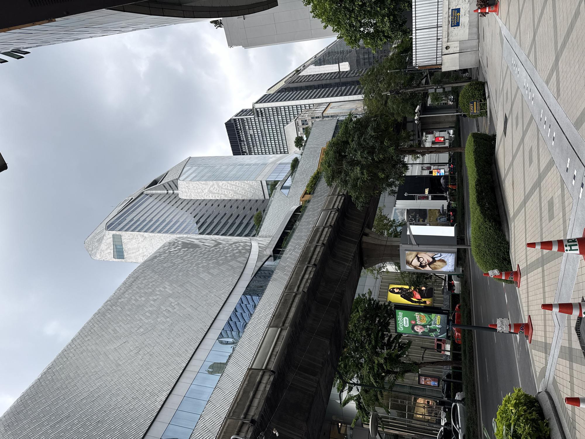 The image depicts a street scene in Bangkok, Thailand. High-rise buildings with modern architectural designs dominate the left side of the frame, their facades primarily composed of glass and light-colored materials. An elevated railway structure runs parallel to the buildings, partially obscuring some of the ground-level structures. Along the street, there are billboards displaying advertisements. The street itself appears to be relatively empty of pedestrian traffic, with a row of orange and white traffic cones placed along the edge. Lush green foliage lines the street, providing a contrast to the concrete and steel of the urban environment. The sky is overcast, suggesting a cloudy day.
